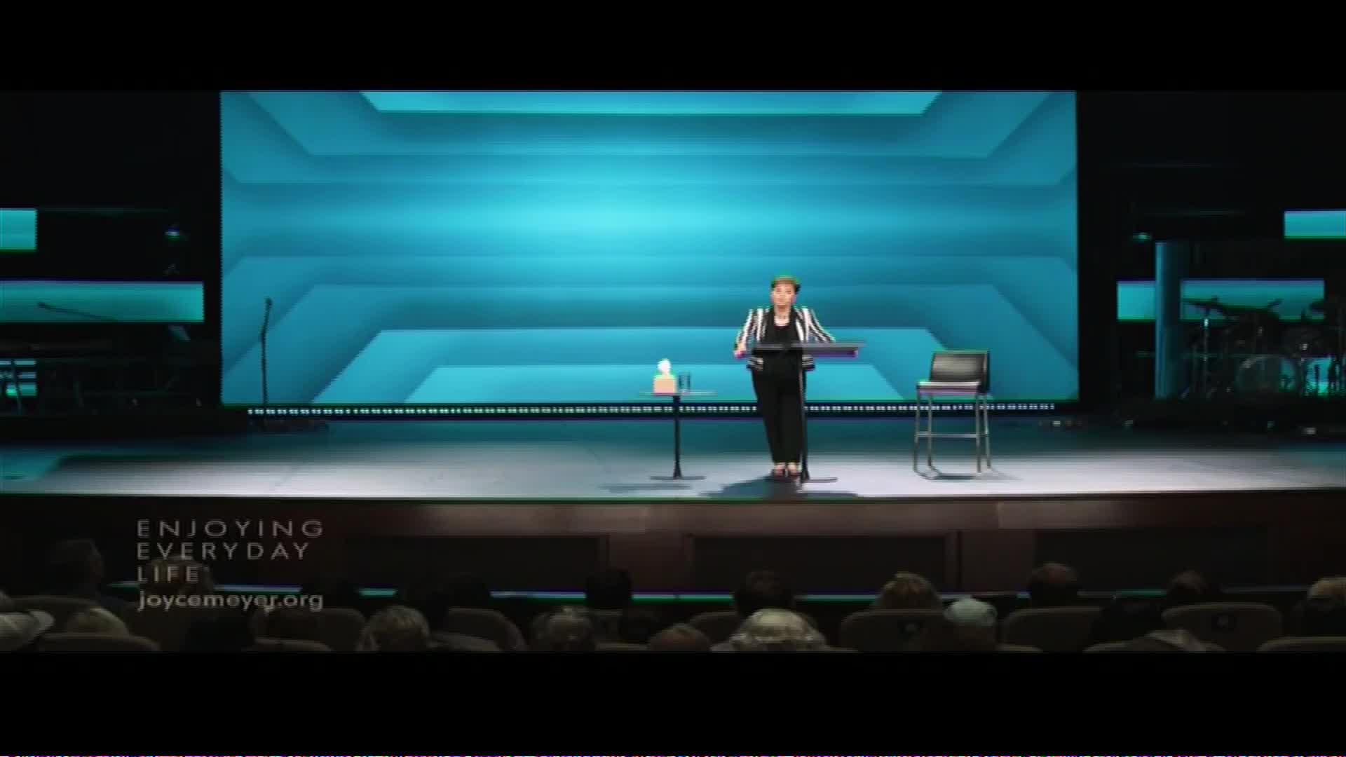 A woman stands at a podium on a stage, speaking to an audience. The backdrop behind her is a large screen displaying a geometric pattern in shades of blue.