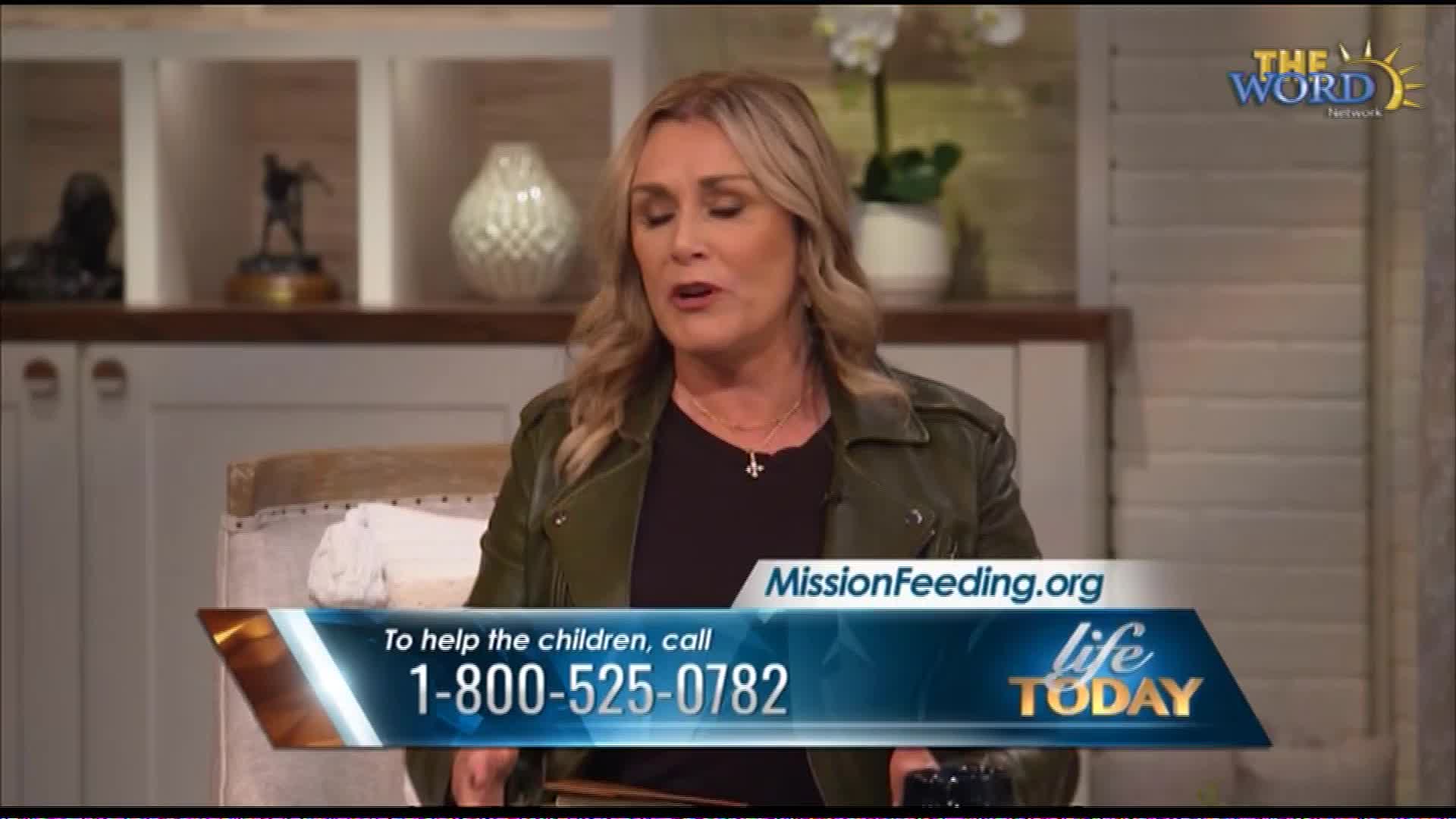 A woman speaks on a set for The Word Network, with a graphic displaying "MissionFeeding.org" and a toll-free number for "Life Today." The background features shelves with decorative items and a plant.