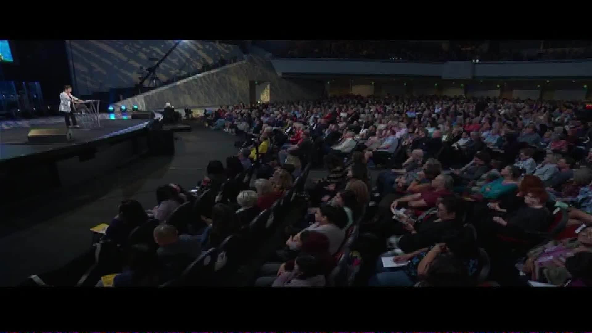 A speaker stands at a podium on a stage, addressing a large congregation gathered in a stadium. The crowd, filling rows of seats, turns their attention toward the speaker.