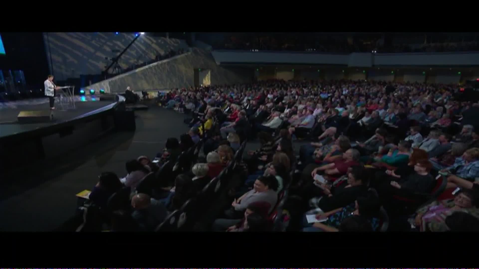 A woman stands at a podium on a stage, addressing a vast audience seated in a stadium. The crowd fills the tiered seating, many looking towards the stage, their faces illuminated by the lights.