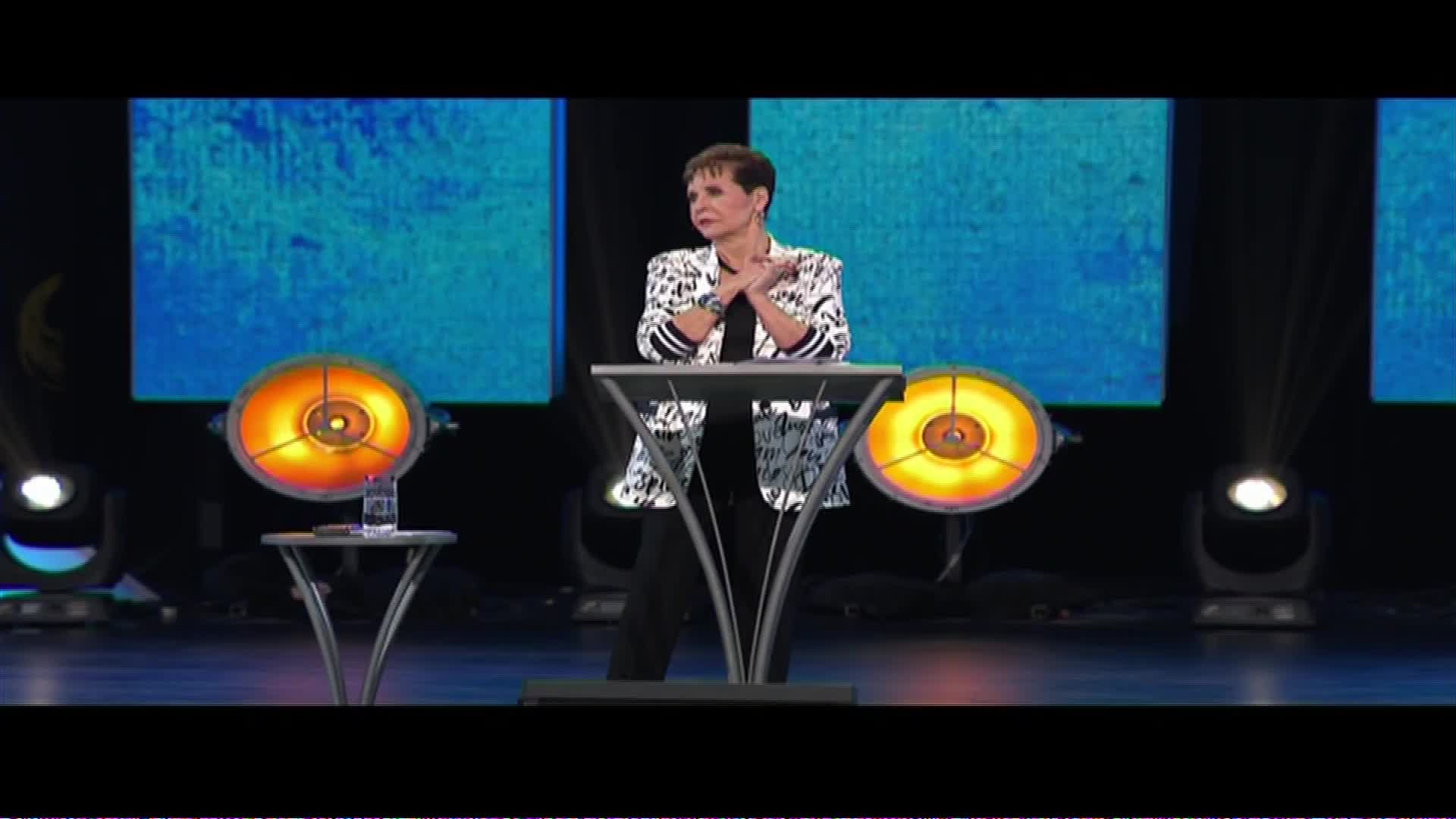 A woman stands behind a podium, her hands clasped. The stage is lit by spotlights and large circular lights, with a blue screen behind her. This is a broadcast from The Word Network, reaching audiences across the United States.