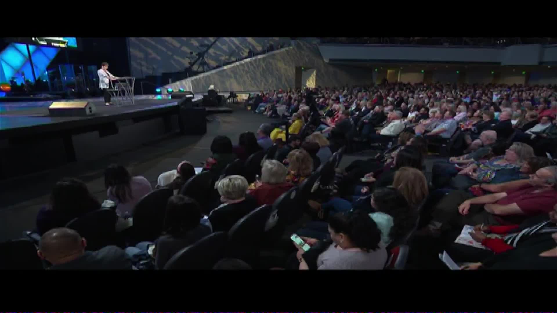 A man stands on a stage, speaking to a large congregation filling the seats before him. The scene is broadcast live across the United States on The Word Network.