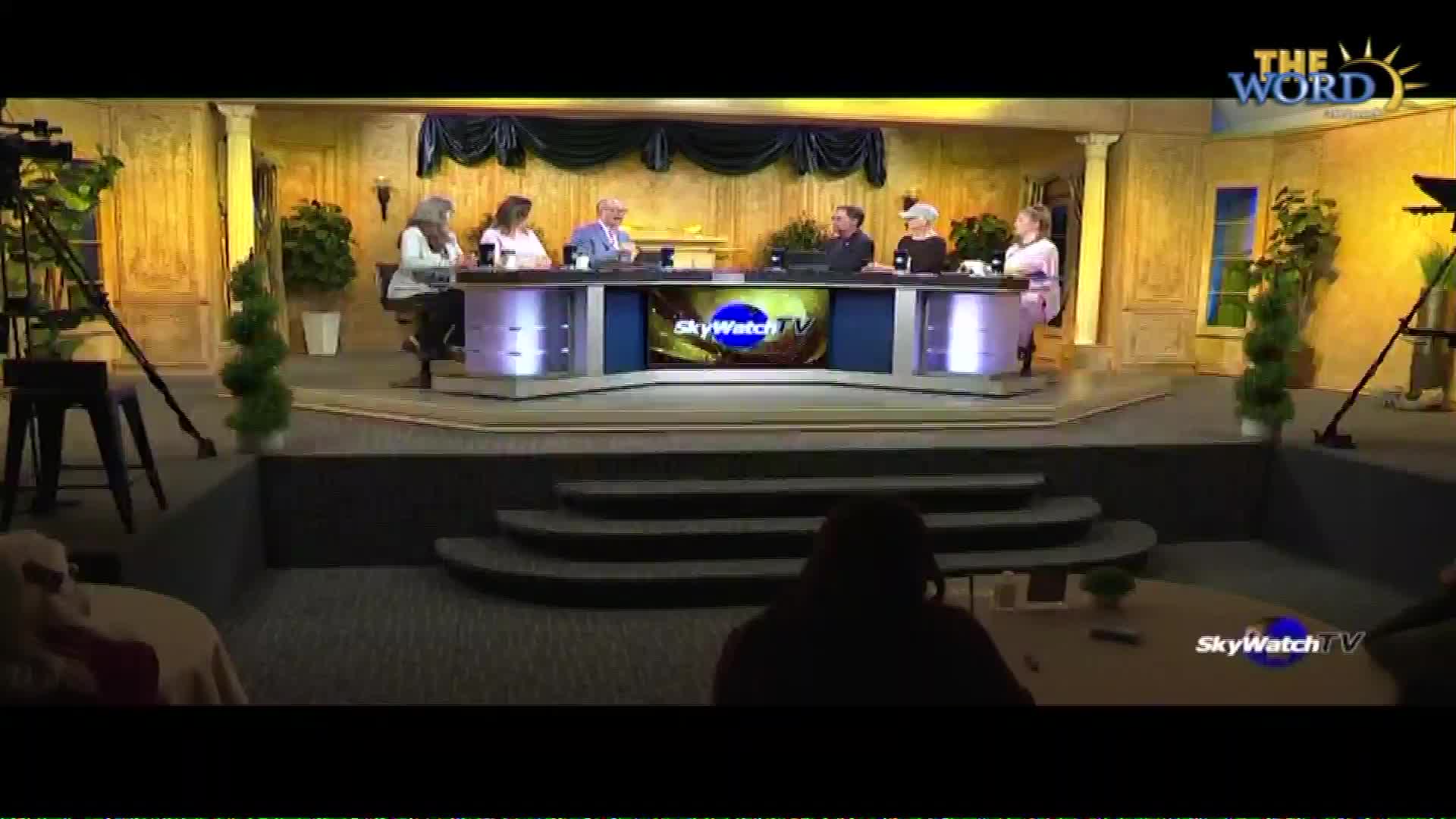 A panel of six people sits behind a desk with "SkyWatch TV" displayed on a screen. Cameras on tripods are positioned on either side of the stage, ready to capture the broadcast.