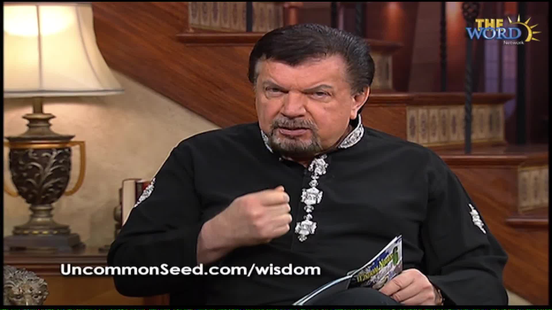 A man in a black robe with ornate silver trim gestures with his right fist as he speaks, holding a magazine in his lap. The Word Network logo is visible in the upper right corner of the screen.