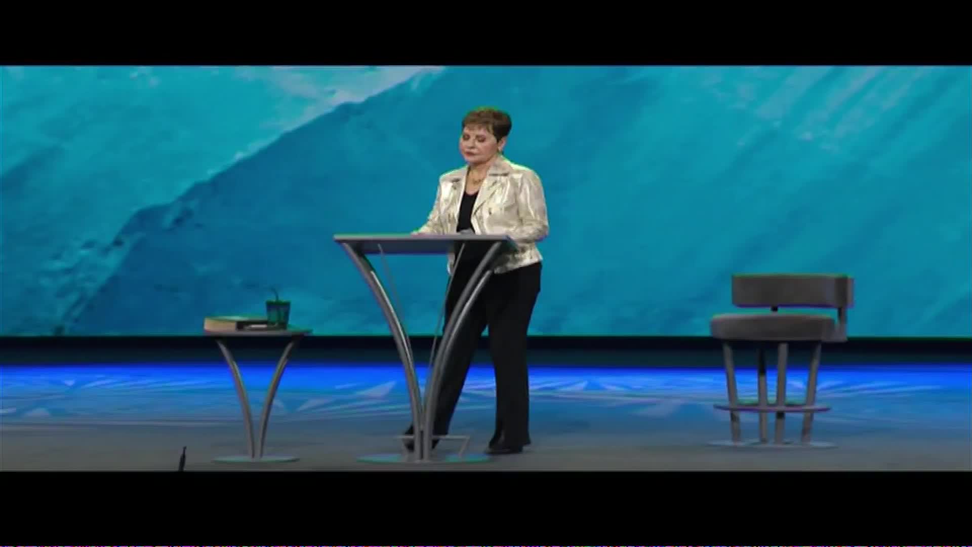 A woman stands behind a podium, her hands resting on its surface. She's dressed in a shimmering jacket and dark pants, facing a large blue backdrop.
