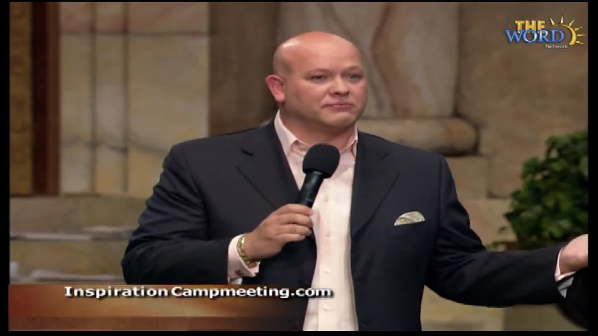 A bald man in a dark suit speaks into a microphone, his left arm extended. The Word Network logo appears in the upper right corner.