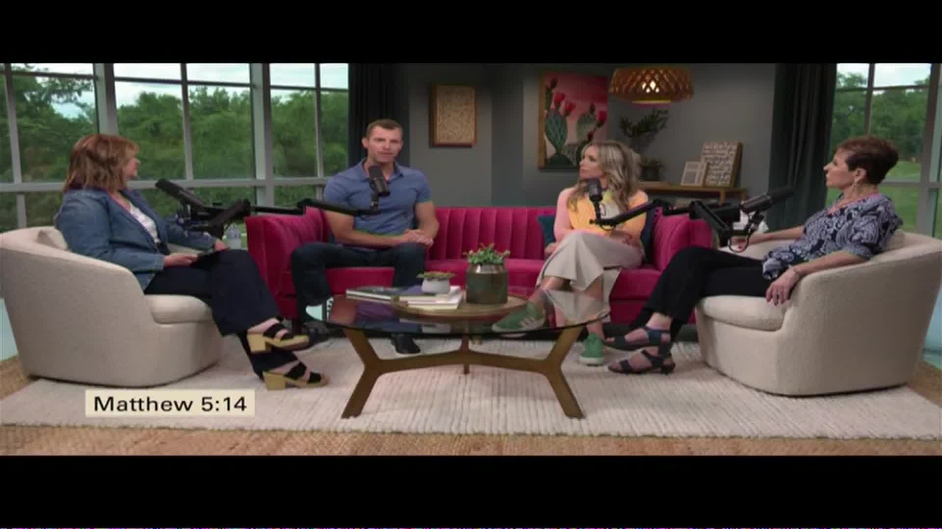 Four people are seated in a studio, with a bright pink couch and plush armchairs. A glass coffee table sits between them, holding a few books and a small plant. The background features large windows looking out onto lush greenery.