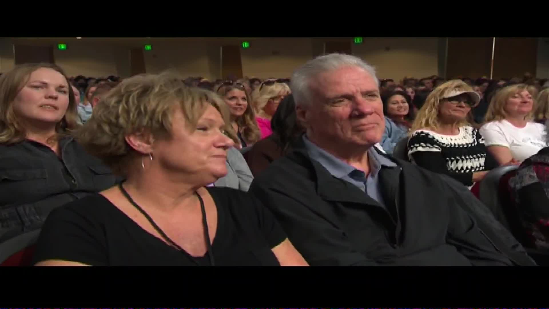 A man and woman sit in the front row of a packed auditorium, their faces turned towards something off-camera. The man's gaze is steady, while the woman's eyes are slightly narrowed, as if listening intently to a speaker at The Word Network event.