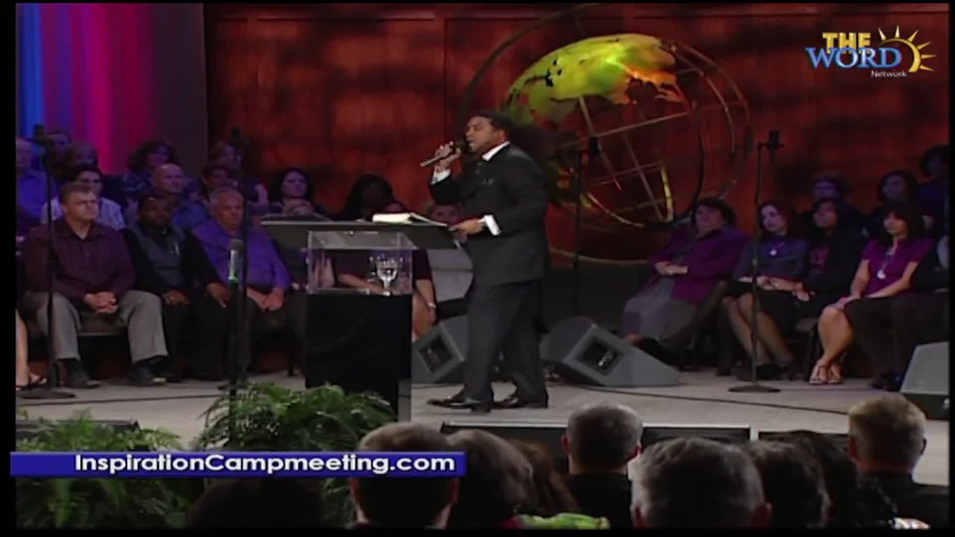 A man in a suit speaks into a microphone from behind a podium, holding a book. The Word Network logo appears in the upper right corner of the screen, and a banner at the bottom reads "InspirationCampmeeting.com."