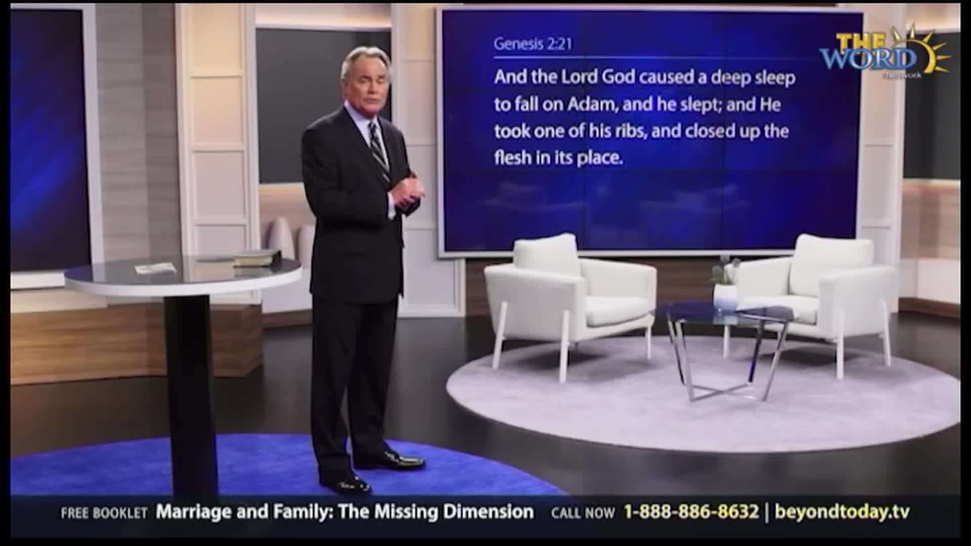 A man in a dark suit stands on a blue rug in a studio. Behind him, a large screen displays text from Genesis, with "The Word Network" logo in the corner.
