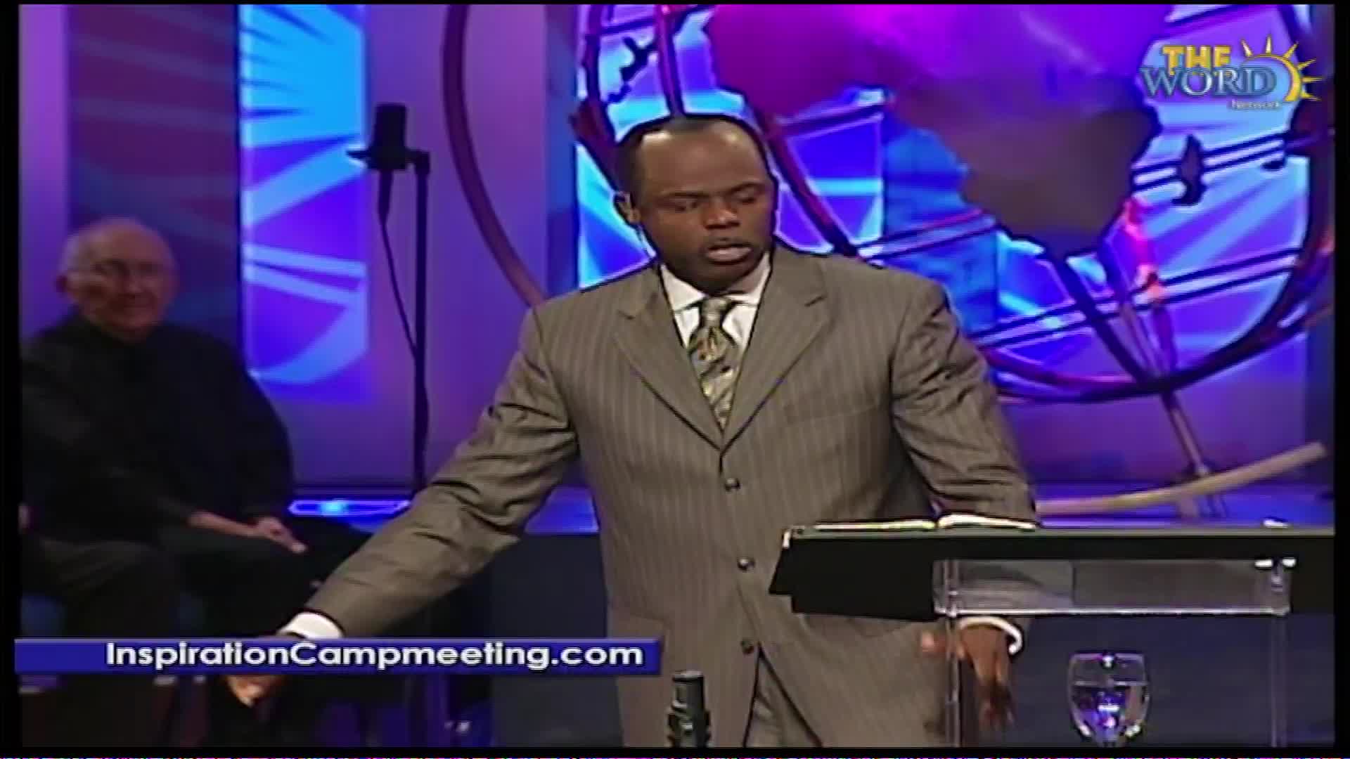 A man in a pinstripe suit stands at a podium, speaking. Behind him, an older gentleman sits in a chair, and a large globe-like structure is visible. The Word Network logo appears in the upper right corner.