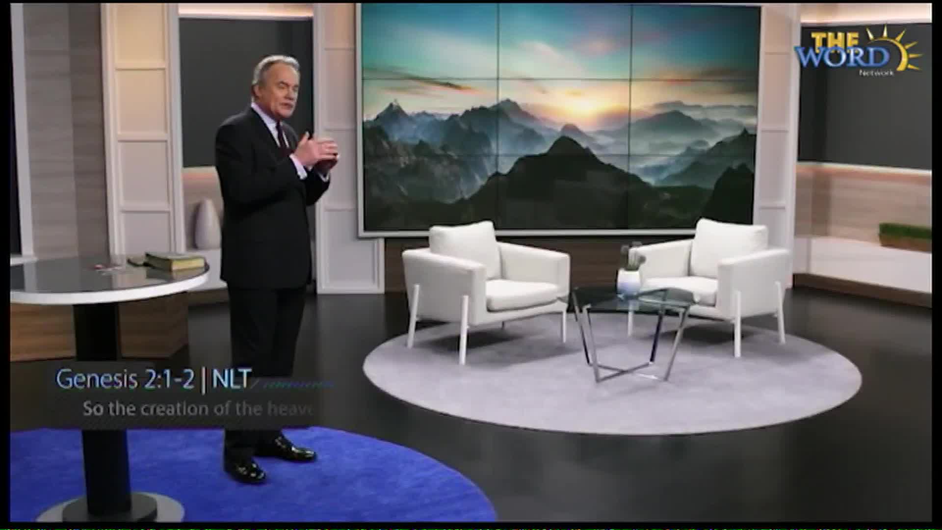 A man in a suit stands on a blue rug, gesturing with his hands. Behind him, a large screen displays a majestic mountain landscape bathed in sunrise light. The Word Network logo is visible in the upper right corner.