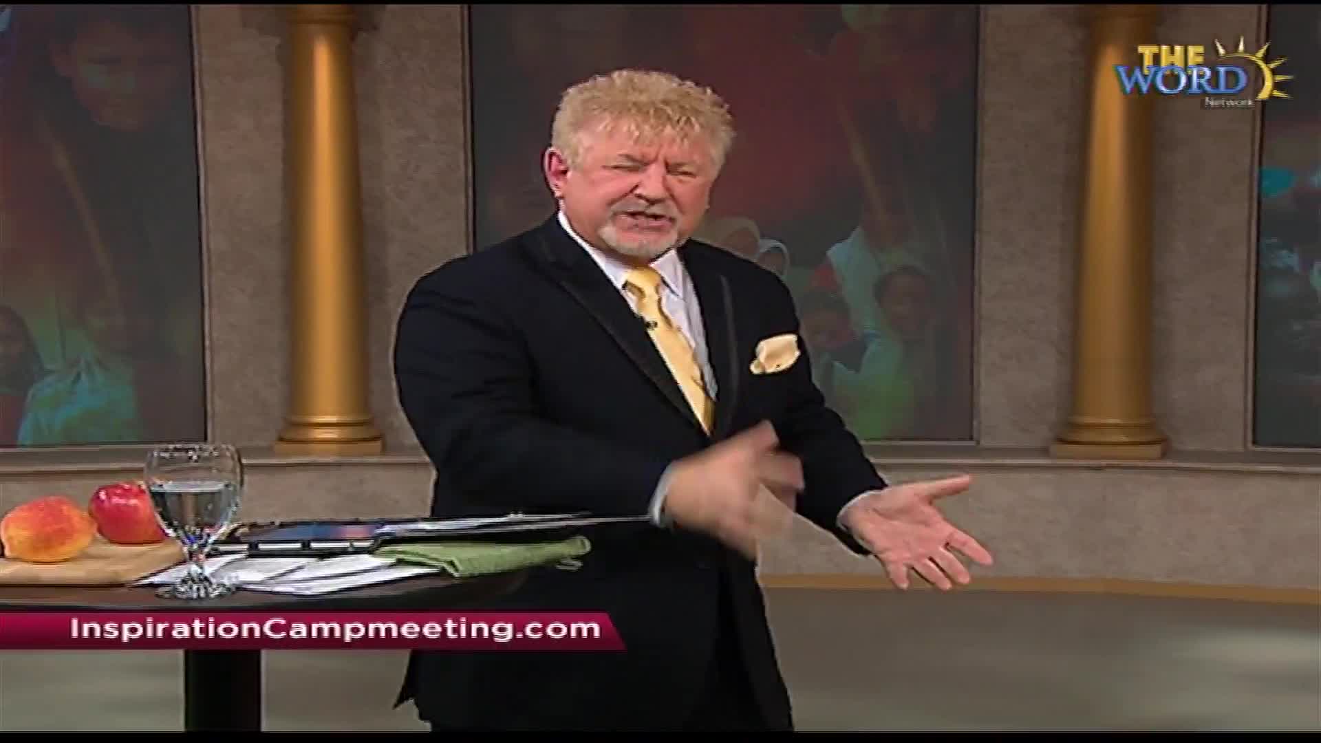 A man in a black suit and yellow tie gestures with his hands while speaking. He's on a set with golden columns and a screen showing people.