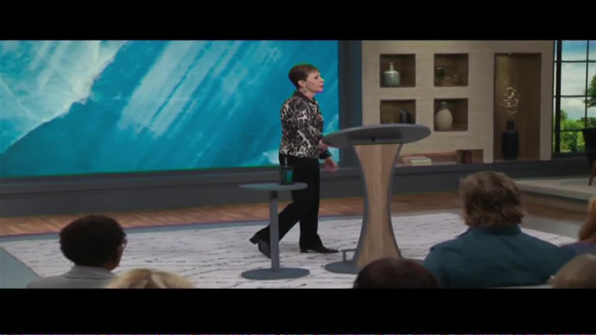 A woman in a patterned jacket walks across a stage, heading towards a podium. The Word Network broadcast plays behind her, featuring a bright blue, abstract background.