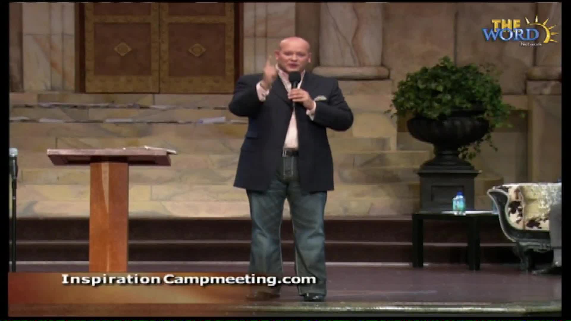 A man in a dark suit jacket and jeans holds a microphone and gestures with his right hand. He stands on a stage with a wooden podium to his left. The Word Network logo is visible in the upper right corner.