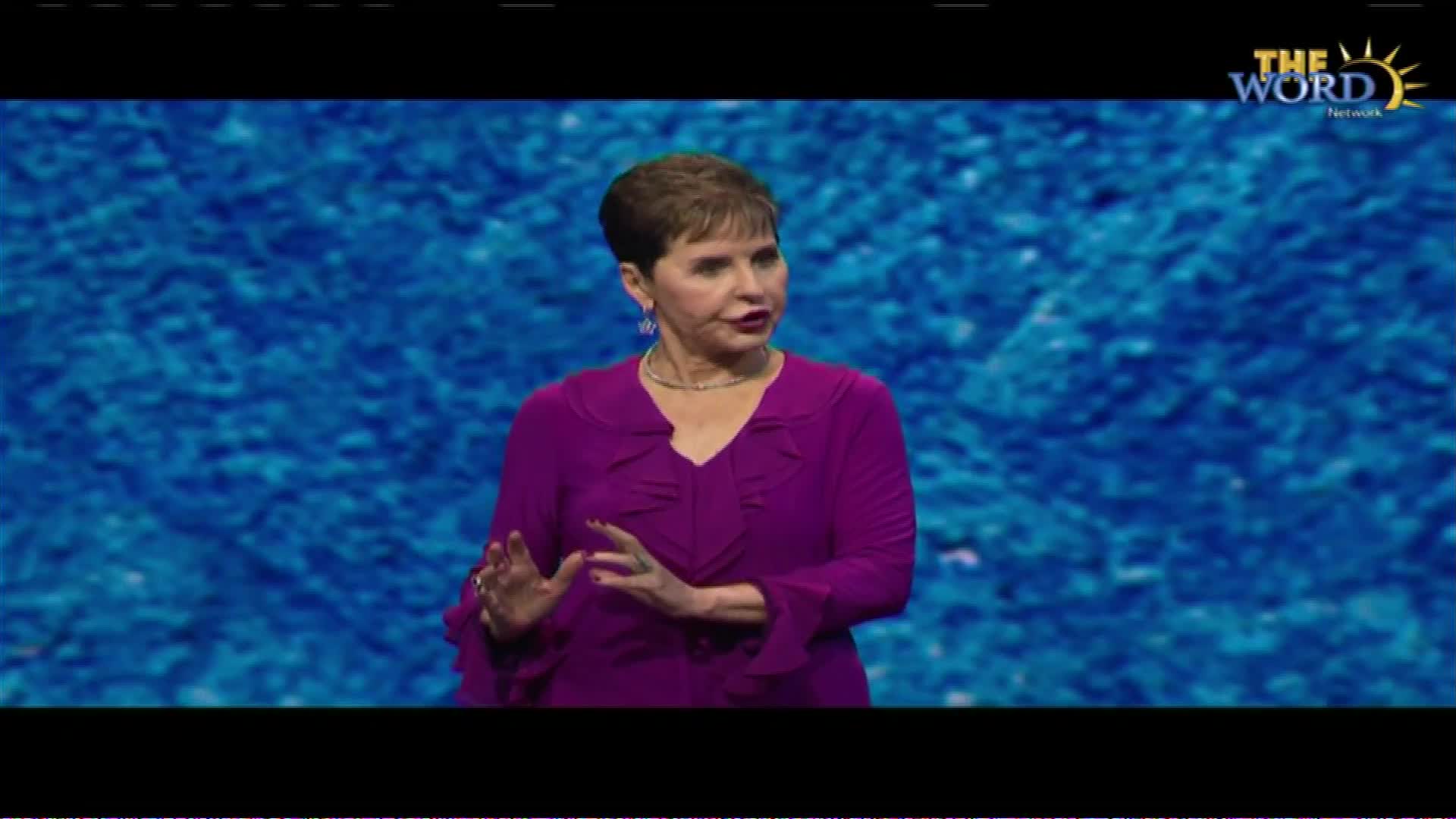 A woman in a vibrant purple top stands before a textured blue backdrop. She gestures with her hands as she speaks, her expression engaged. The Word Network logo is visible in the upper right corner.