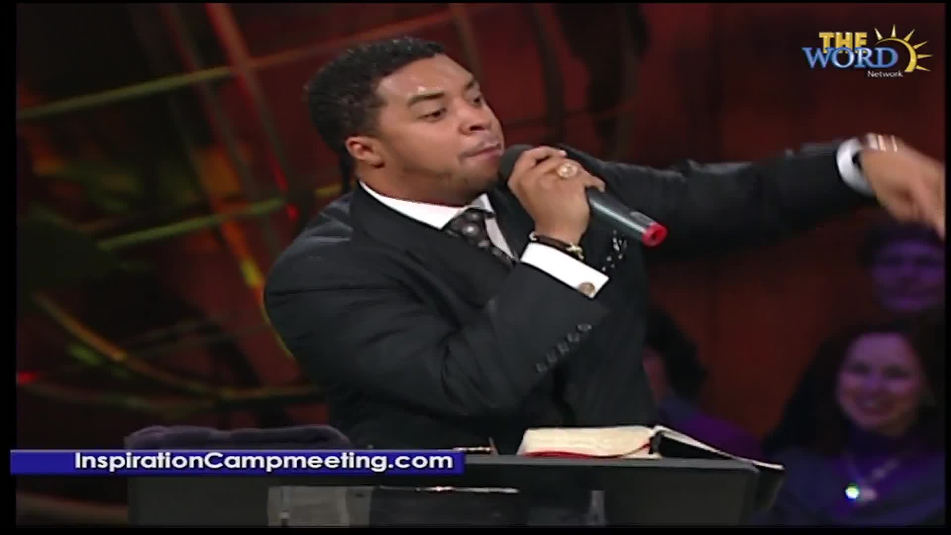 A man in a suit speaks into a microphone, gesturing with his free hand. The Word Network logo is visible behind him.