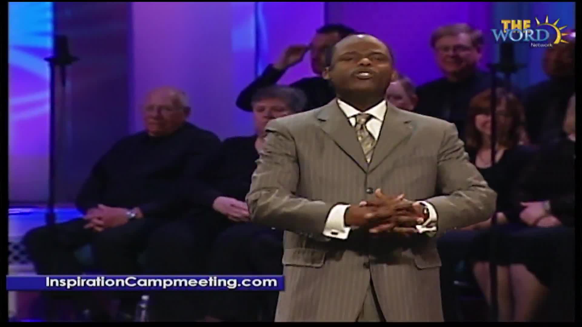 A man in a pinstripe suit stands with his hands clasped, speaking to the audience. Behind him, a choir is gathered, bathed in purple stage lights, as seen on The Word Network.