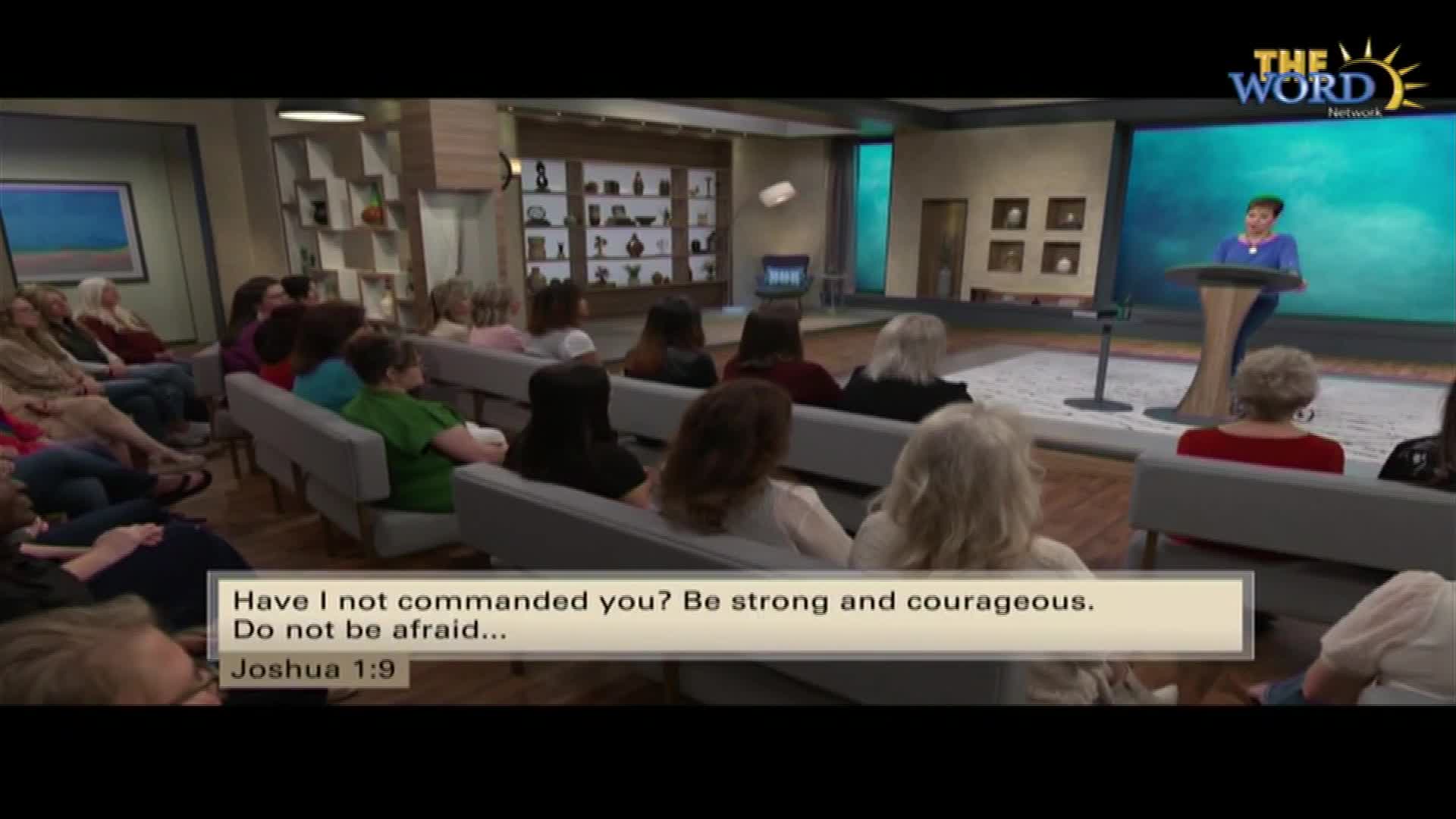 A woman stands at a podium on a stage, speaking to an audience seated in rows. The Word Network logo appears in the upper right corner.