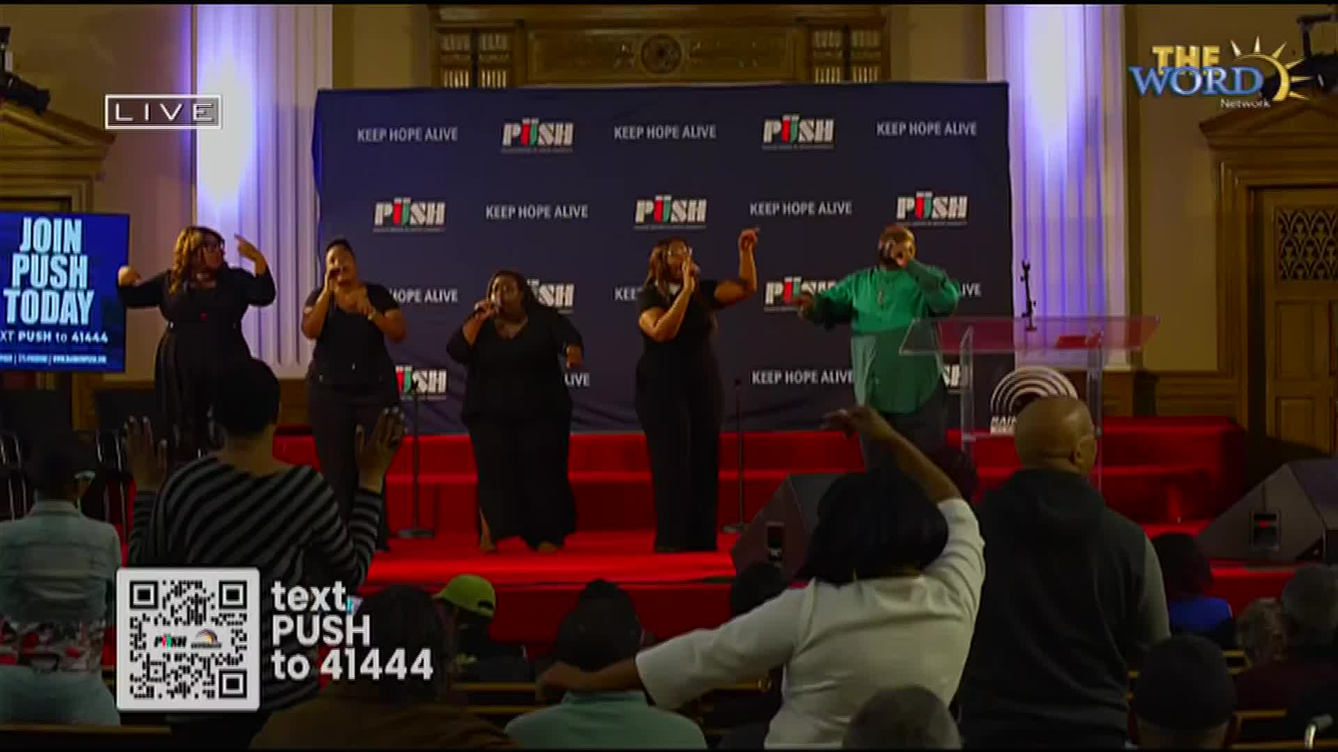 A group of singers, dressed in black, are on stage with microphones, their voices lifted in song. One singer in a bright green shirt stands at a podium, also singing.