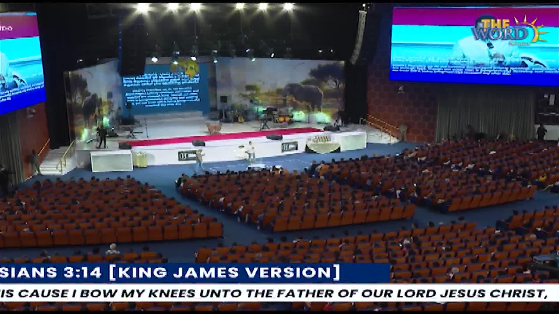 A speaker addresses a large congregation from a raised platform, while a screen behind them displays "THE WORD" logo. Rows of people fill the auditorium, facing the stage.