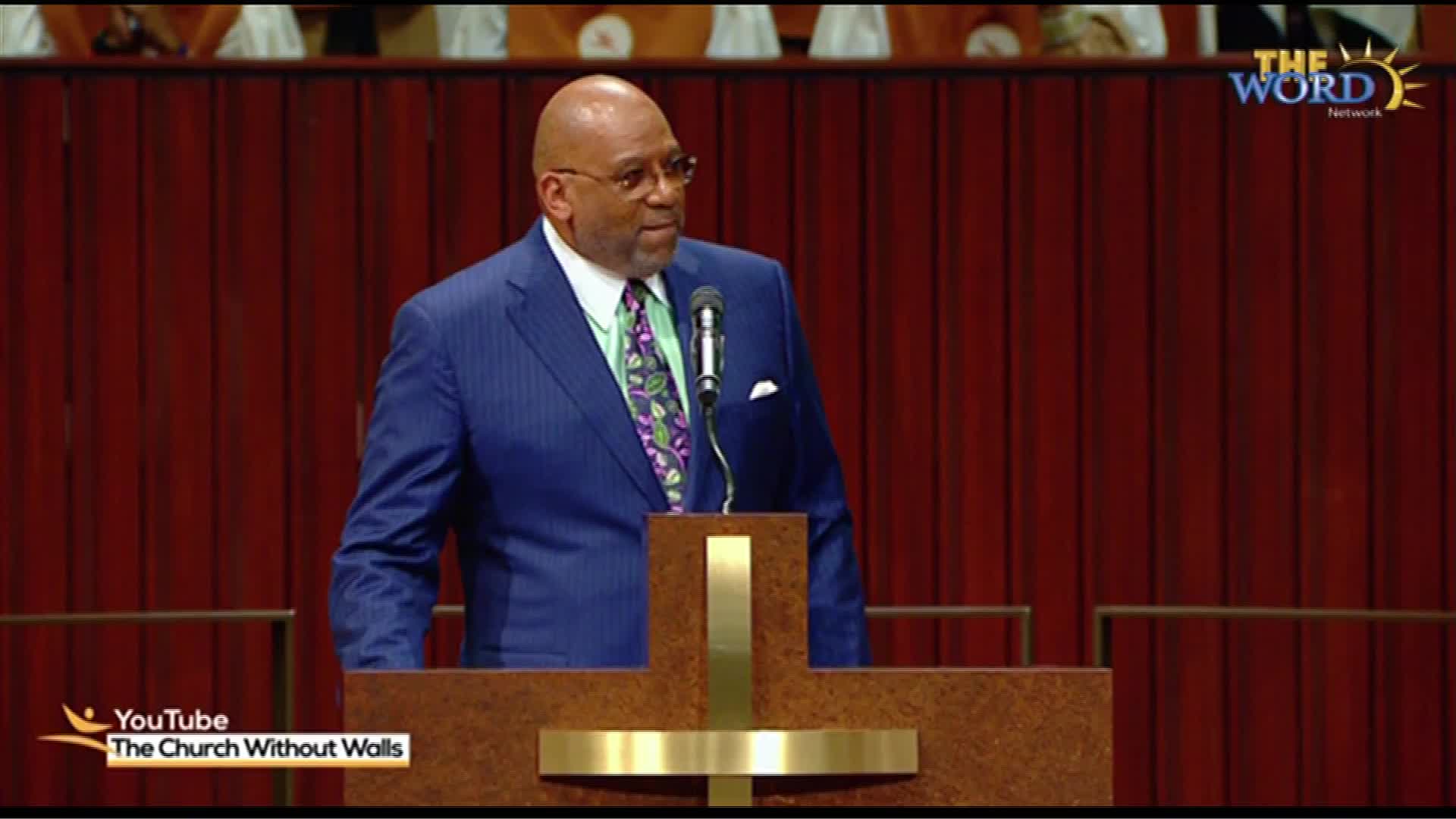 A man in a blue pinstripe suit stands at a wooden pulpit, speaking into a microphone. The backdrop is a rich, dark wood paneling, and a logo for "The Word Network" is visible in the upper right corner.