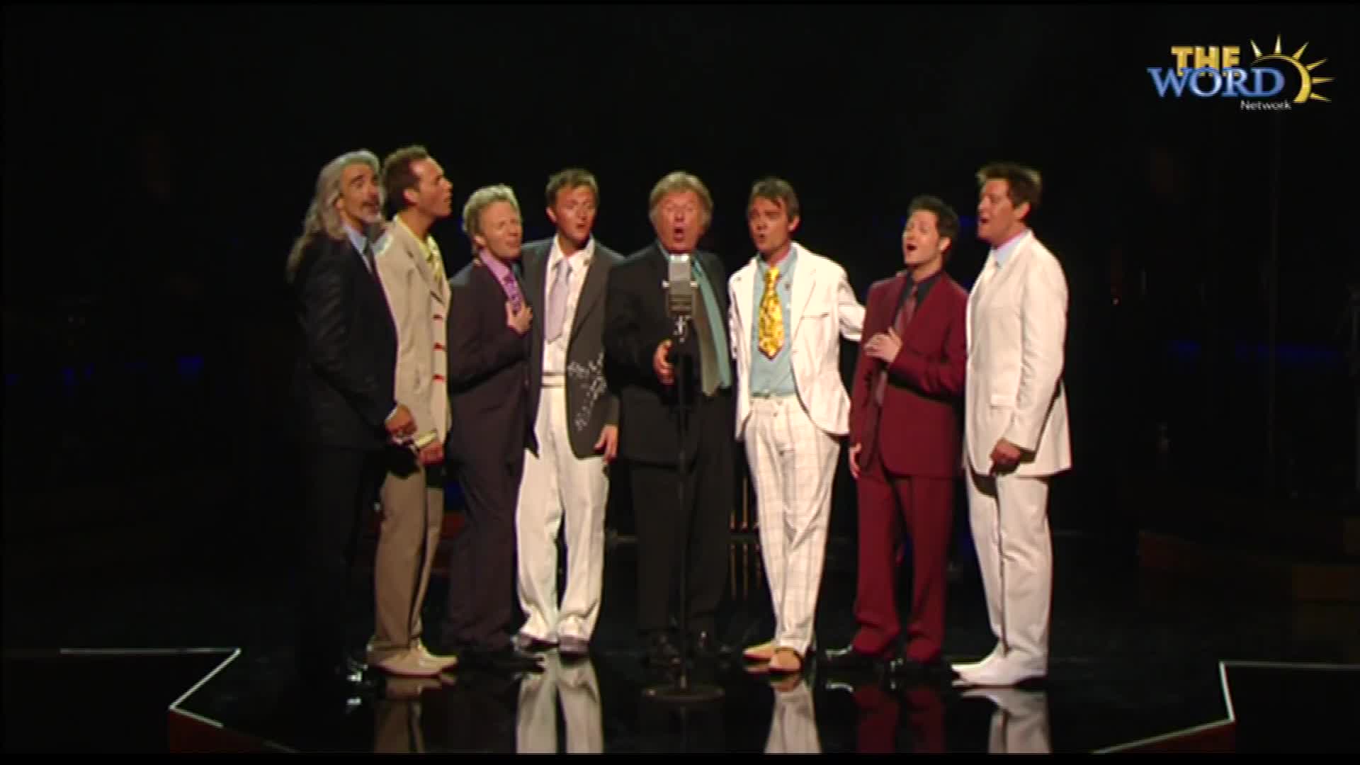 A group of men in suits stand shoulder to shoulder, singing into a single microphone. The Word Network logo is visible in the upper right corner.