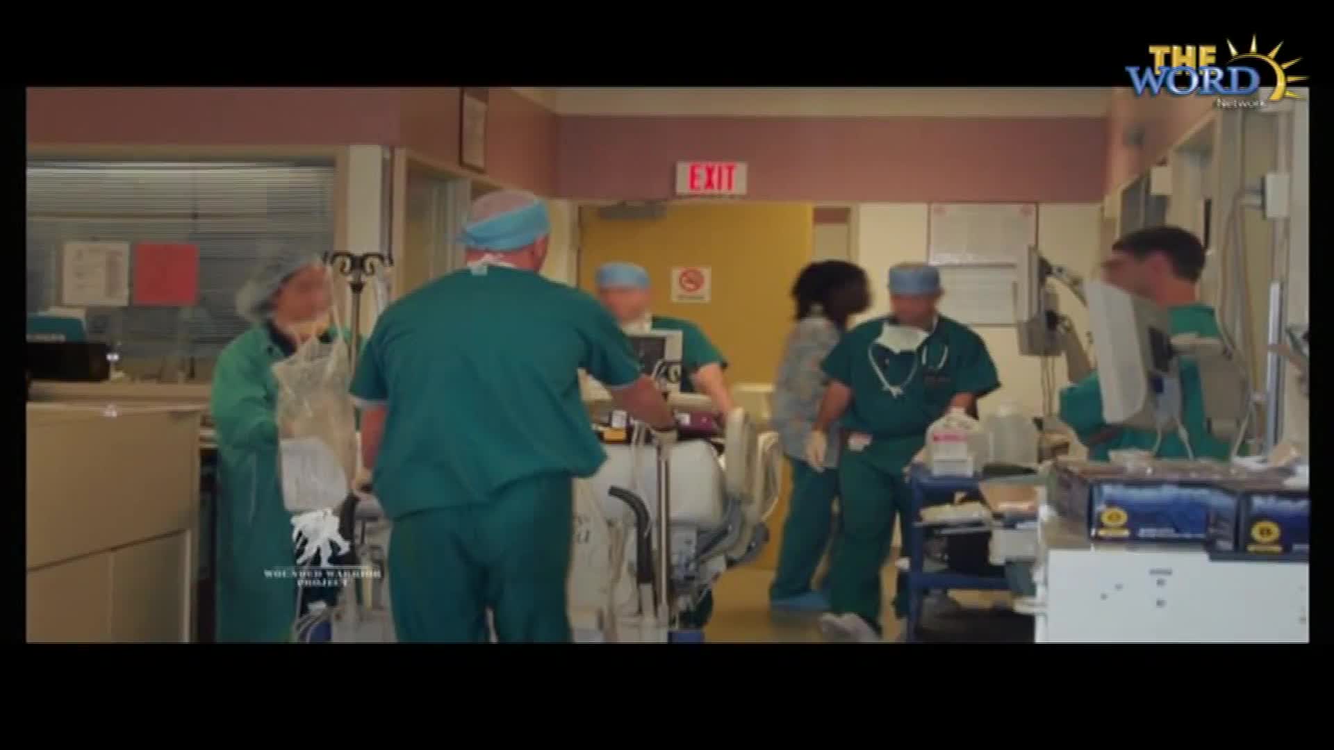 Medical professionals in green scrubs move around equipment in a hospital room. A man in a blue cap adjusts a clear bag.