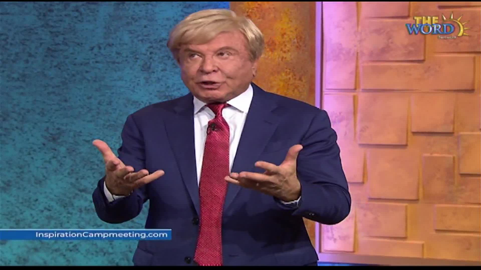 A man in a dark blue suit and red tie gestures with both hands, his blonde hair styled. He speaks with conviction, his voice carrying across the studio.