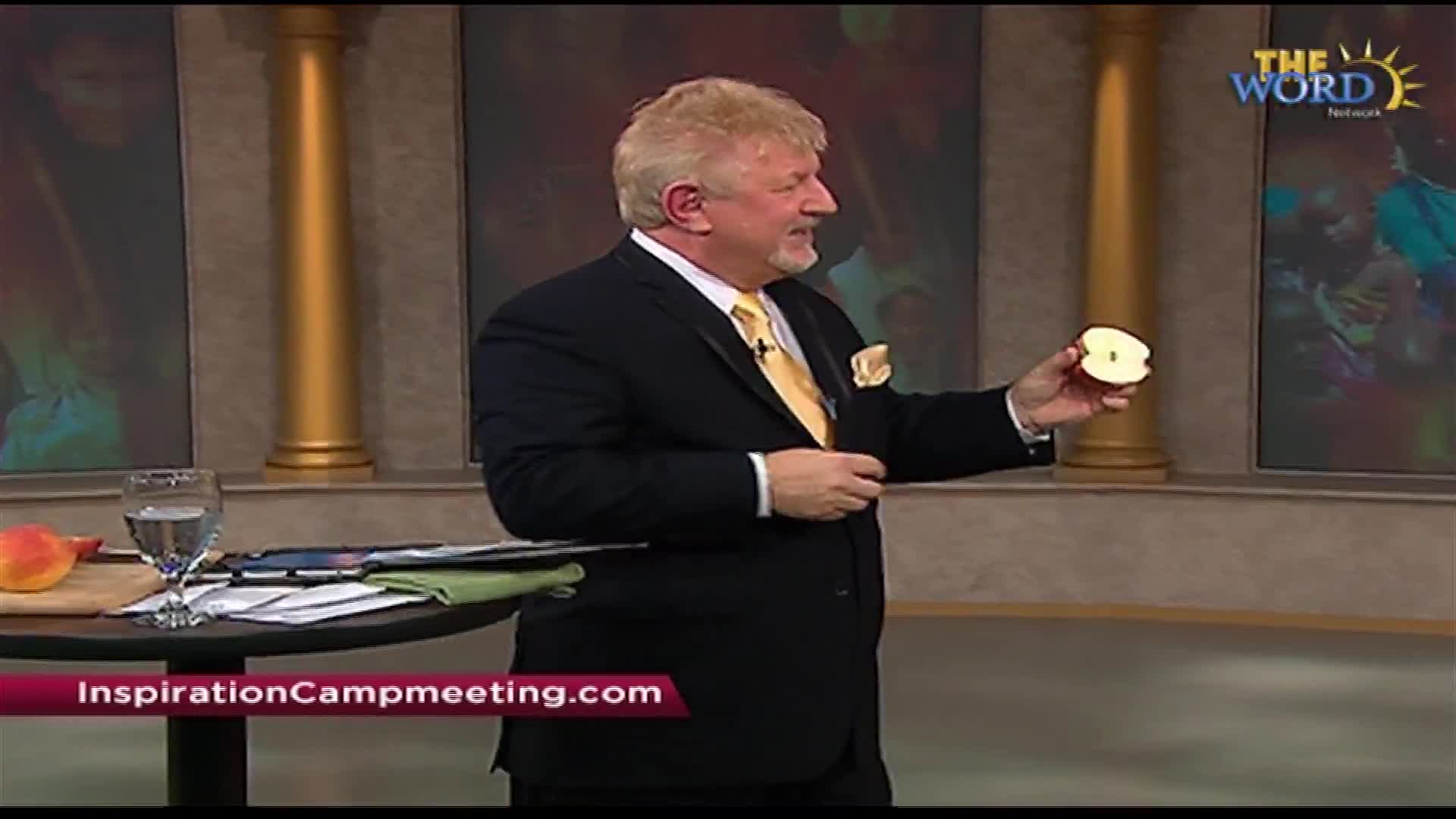 A man in a suit holds up a sliced apple, gesturing with his other hand. On a table beside him sits a whole apple and a glass of water.