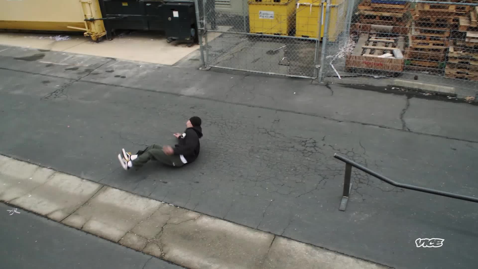 The skater is on his back, sliding across the asphalt after a failed attempt. He's wearing a black hoodie and green pants, his white sneakers kicking up as he tumbles.
