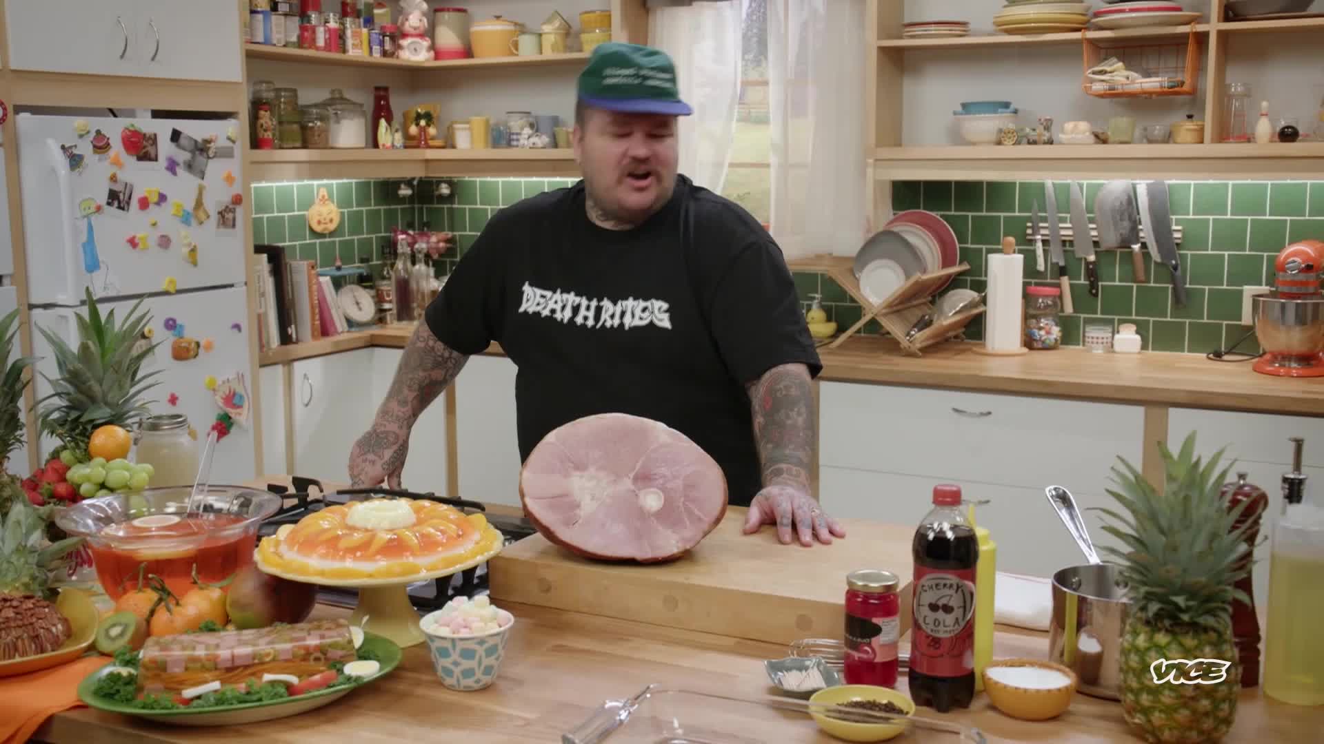 A man in a "Death Rides" t-shirt stands behind a kitchen counter, gesturing toward a massive ham. Various dishes, including a gelatin mold, are arranged on the counter in front of him, as if ready for a feast.
