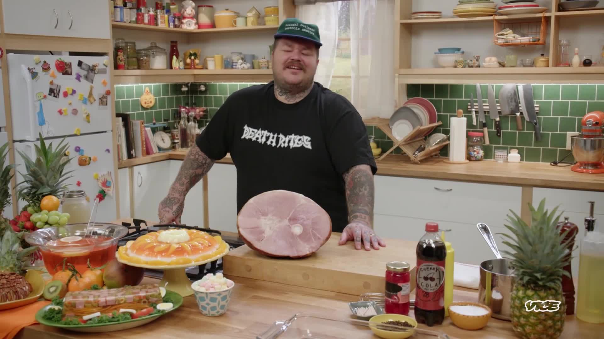 A man with tattoos stands in a kitchen, grinning at the camera. A large ham sits on a cutting board in front of him, surrounded by a spread of food, as part of a VICE TV segment.
