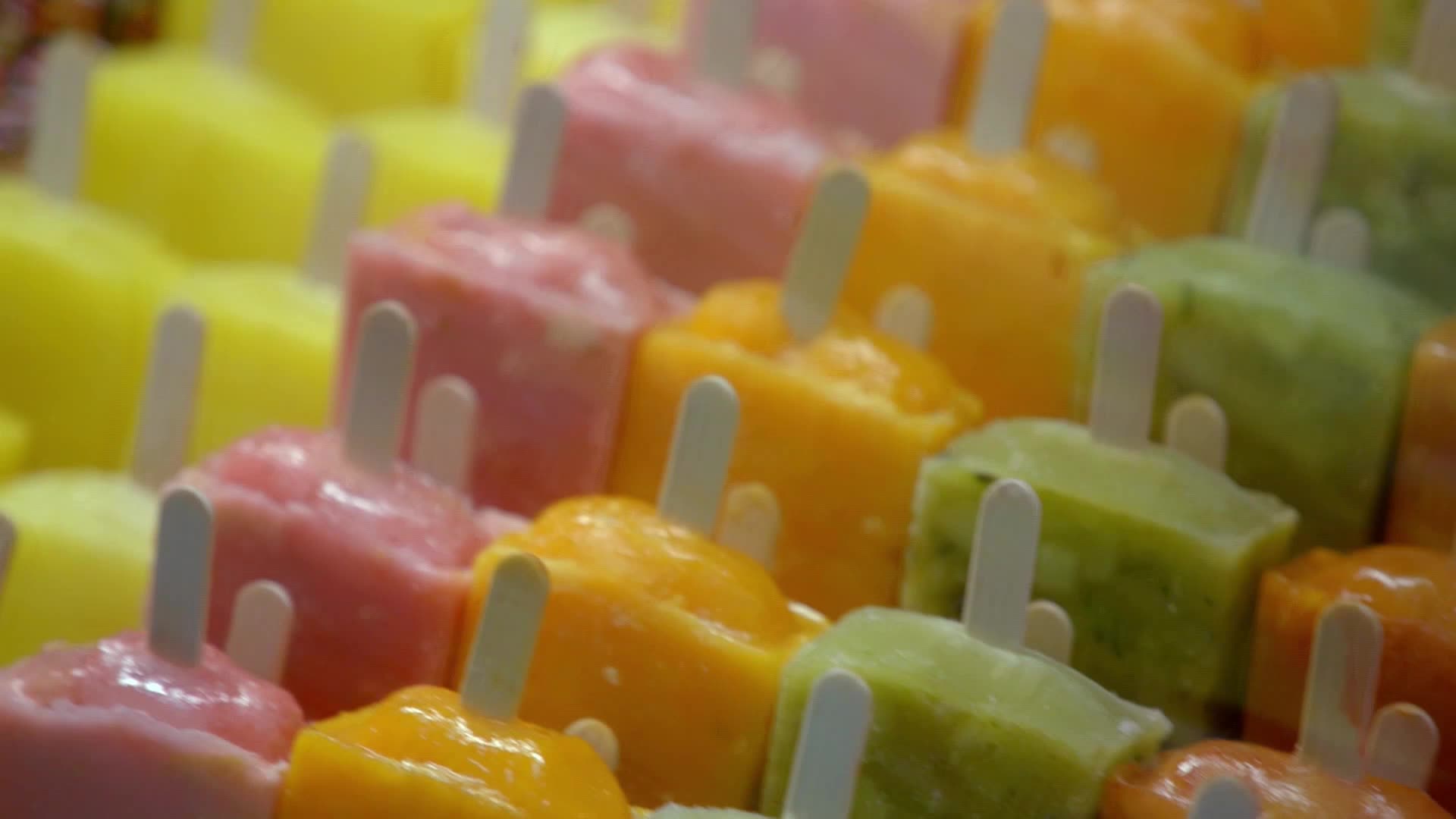 Rows of colorful ice pops, each with a wooden stick, are neatly arranged. The frozen treats, in shades of yellow, pink, orange, and green, are ready to be enjoyed.
