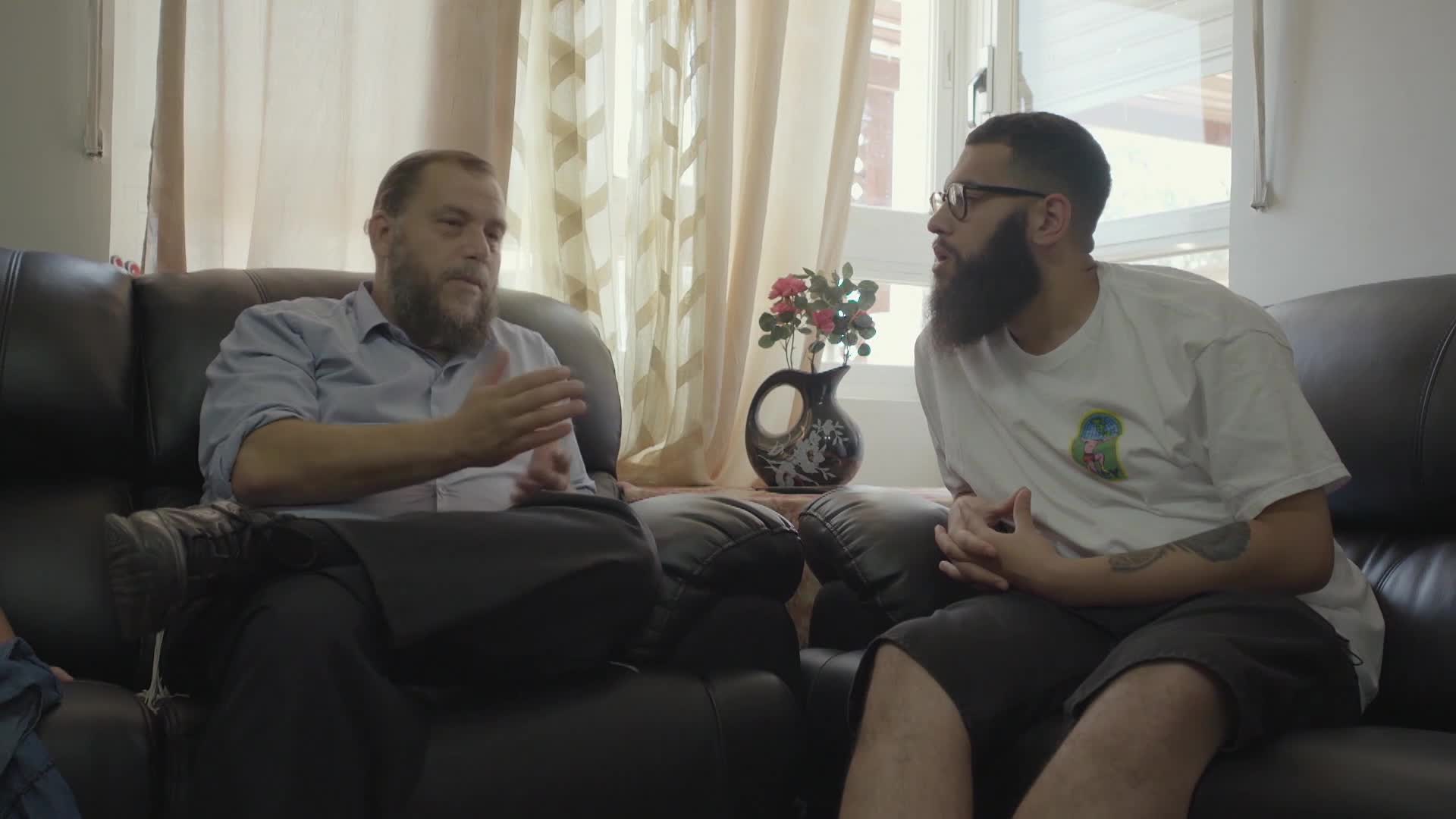 Two men sit on a black leather couch, engaged in conversation. The man on the left gestures with his hands while the other listens intently.
