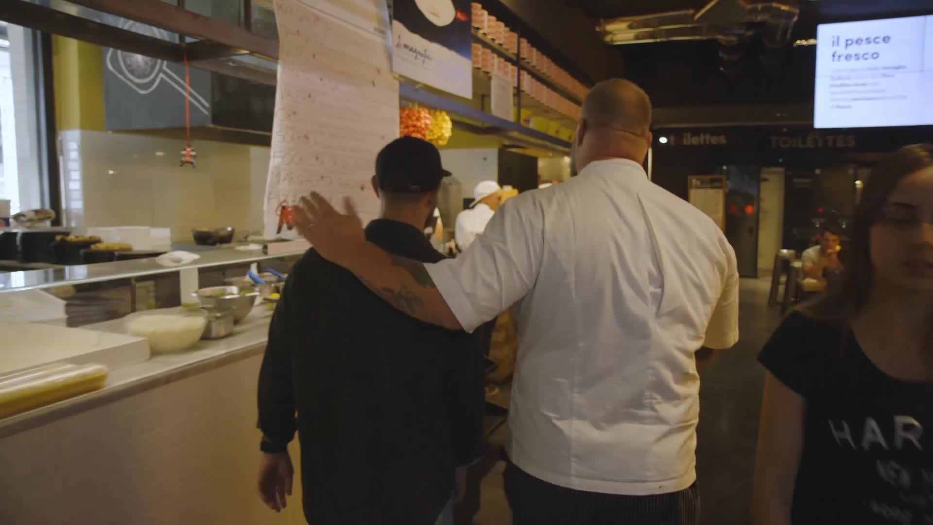 A chef in a white coat has his arm around another man's shoulders, leading him through the restaurant. They're walking past the open kitchen, where a woman is coming toward them.
