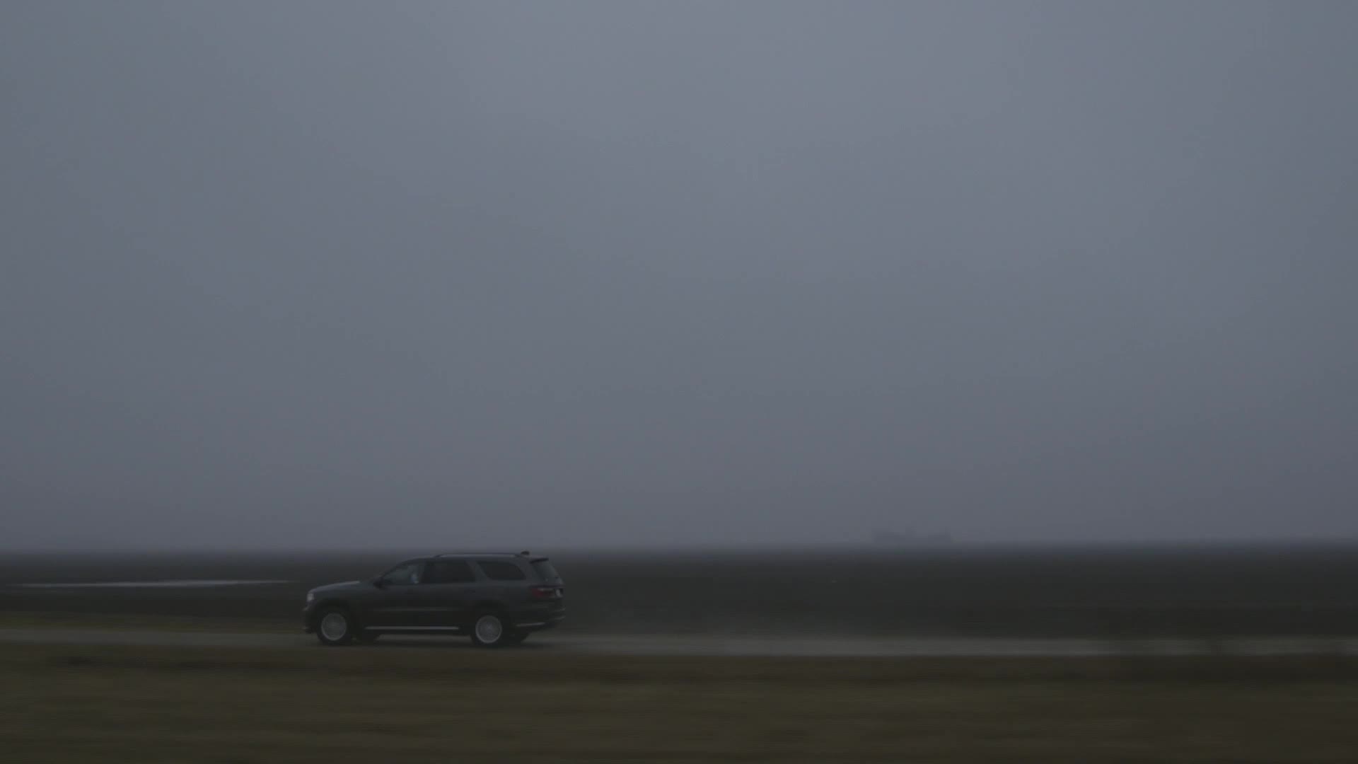 A dark SUV drives along a road, its outline barely visible against the overcast sky. The landscape is flat and shrouded in mist, giving the scene a sense of isolation.
