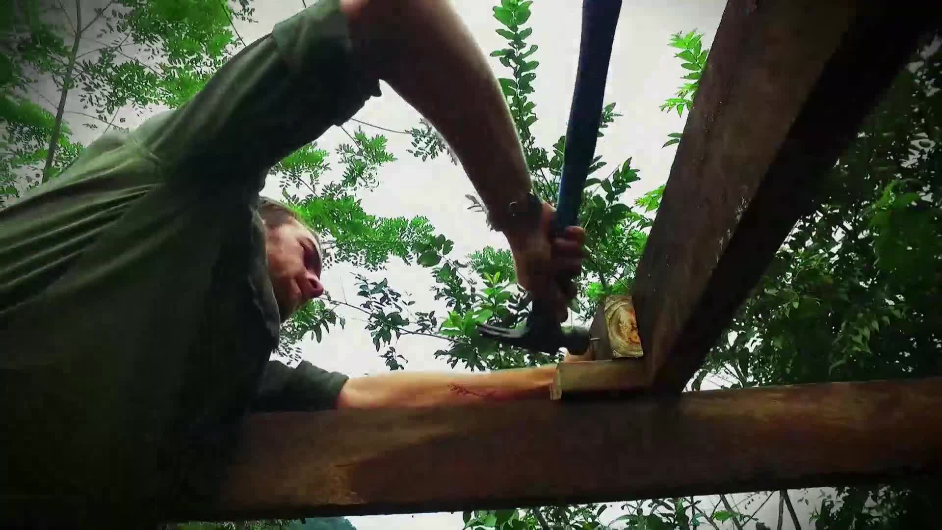 A person in a green shirt is hammering a nail into a wooden beam. The hammer swings down, connecting with the nail head as the wood holds steady.
