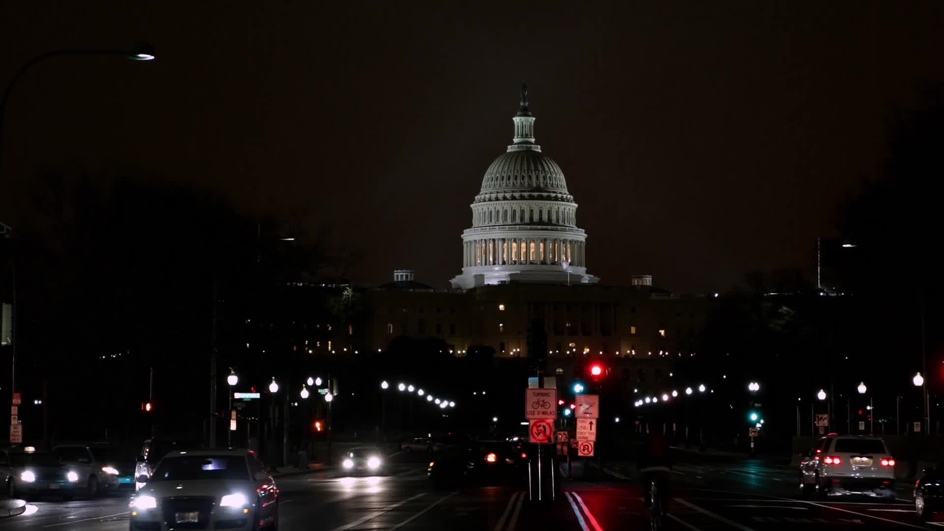 The Capitol building glows under a dark sky, its dome illuminated. Headlights of cars stream down a road, reflecting on the wet asphalt.
