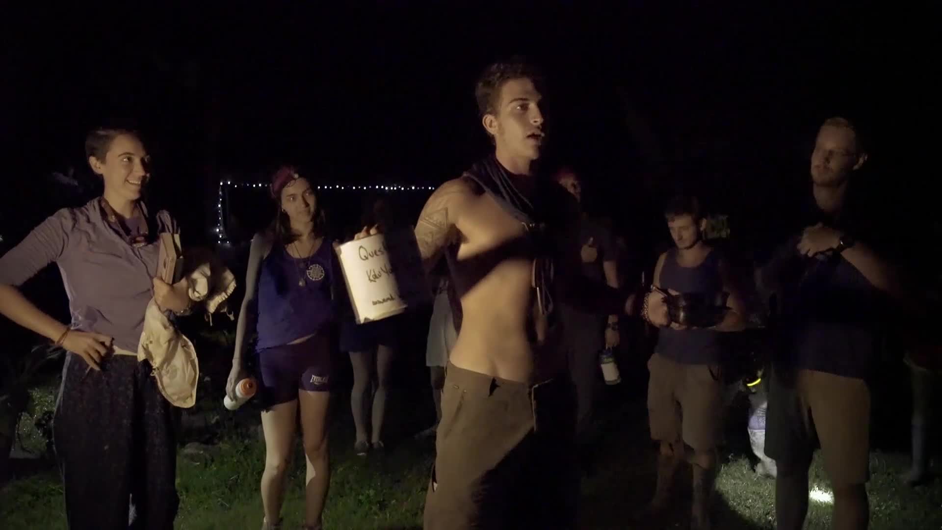 A shirtless man with tattoos gestures while speaking to a group of people gathered at night. Some hold drinks, and one woman clutches a sign that reads "Queer the World."
