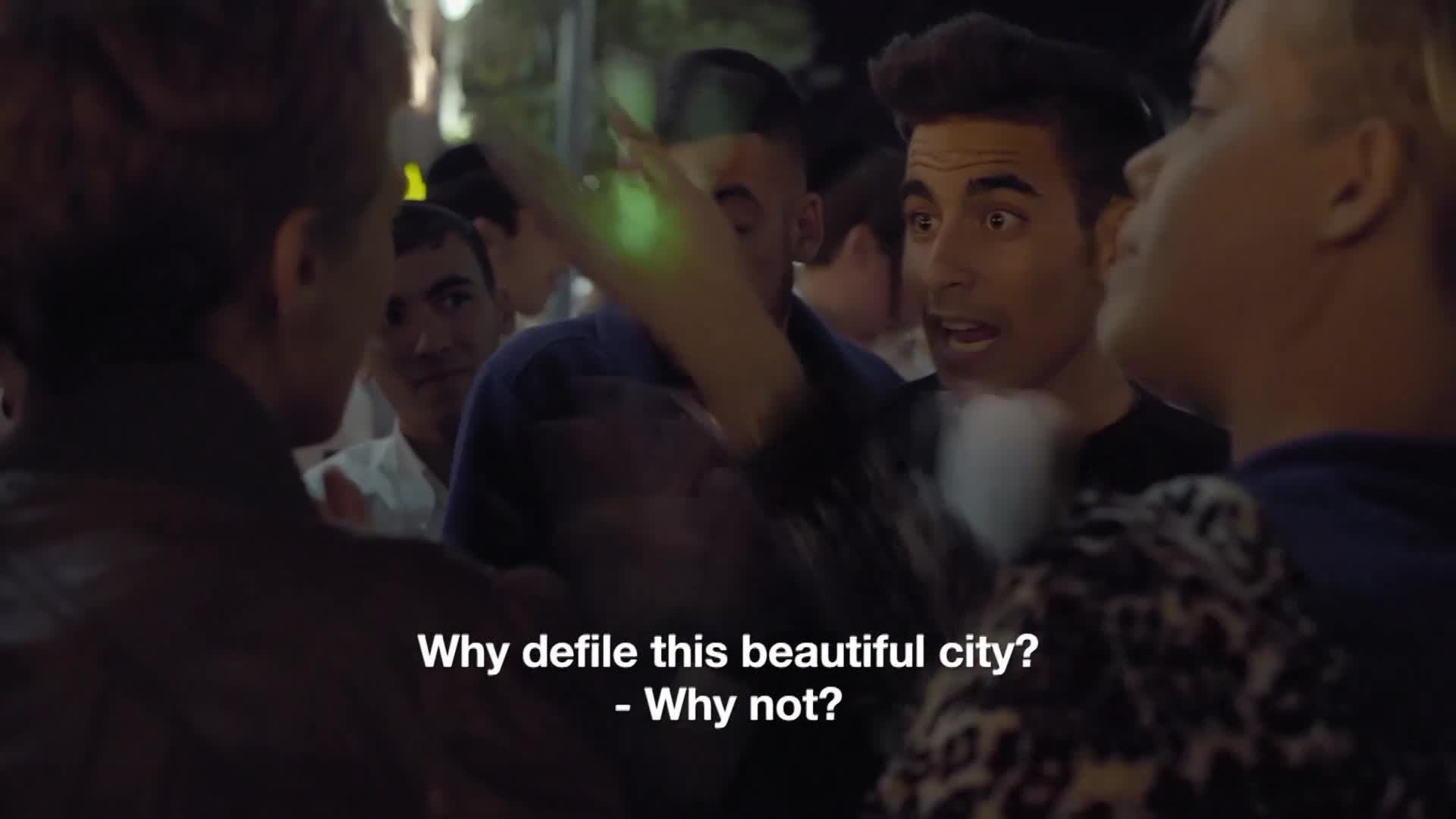 A man with dark hair shouts, eyes wide, while a hand with a glowing green light hovers in the air. Other people surround them, and subtitles on the screen ask, "Why defile this beautiful city?"
