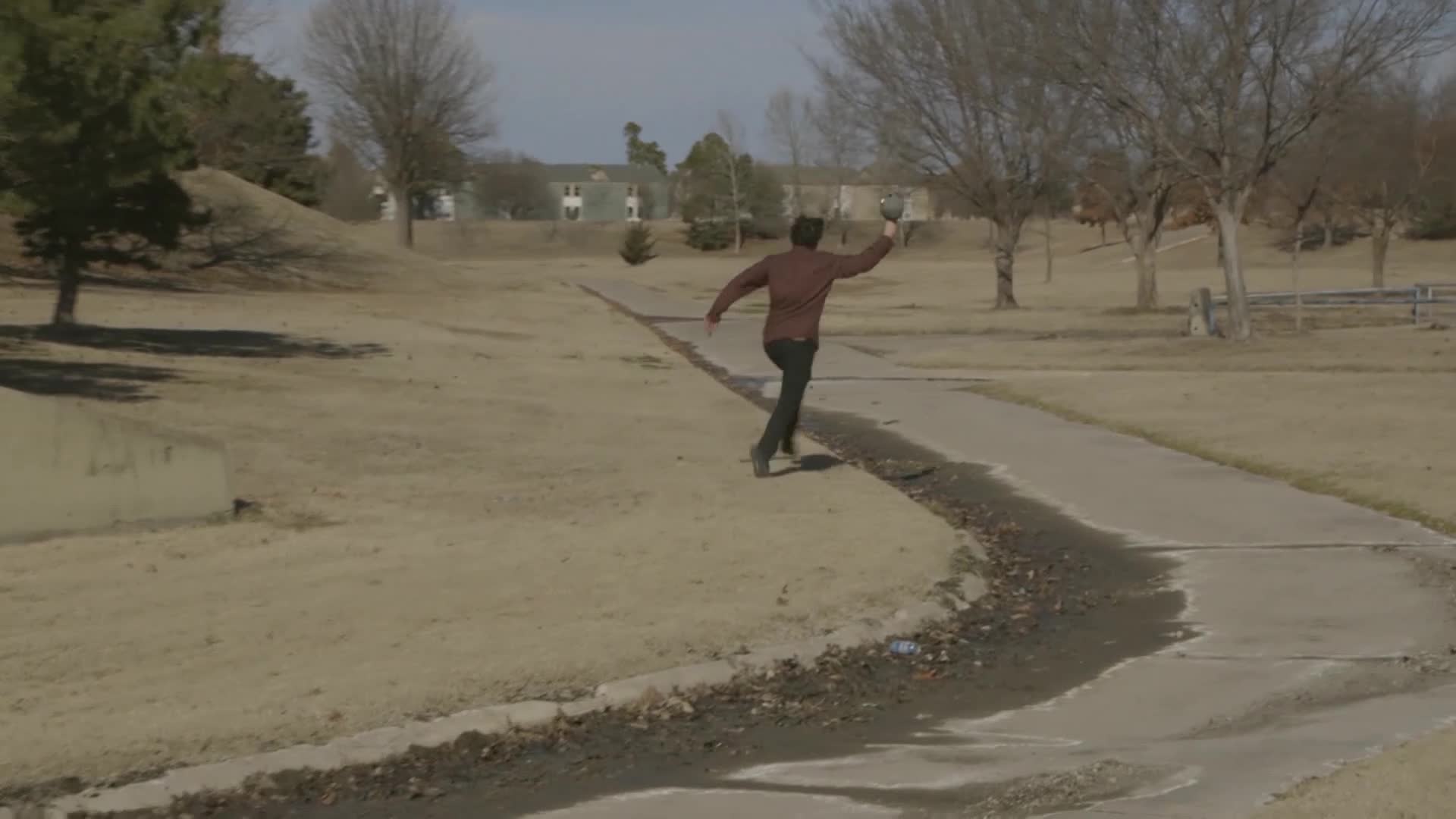 A man in a maroon shirt sprints down a concrete path, holding a small object in his raised hand. He's running away from the camera, toward a cluster of bare trees and a few distant buildings.
