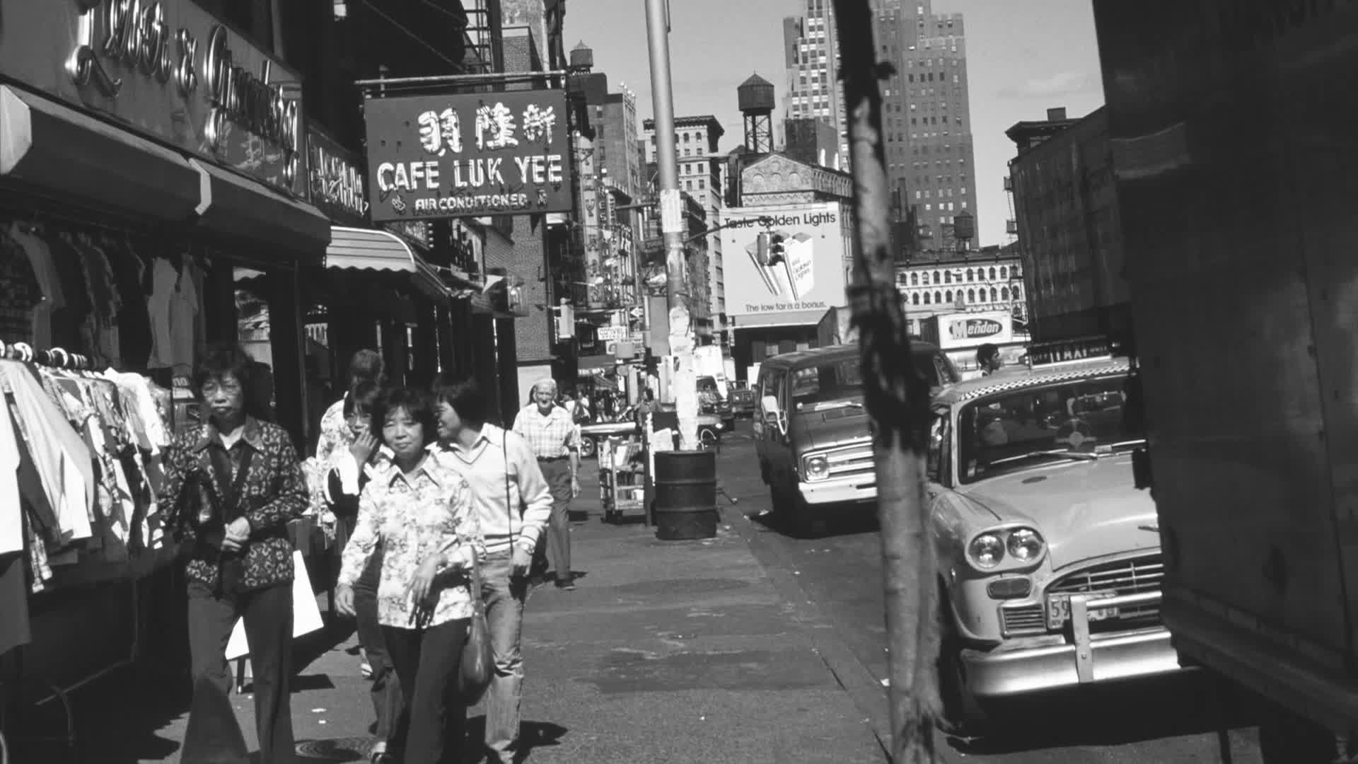 People stroll down a bustling New York City street, passing shops and a parked taxi. The scene is framed by a mix of storefronts and towering buildings under a bright sky.
