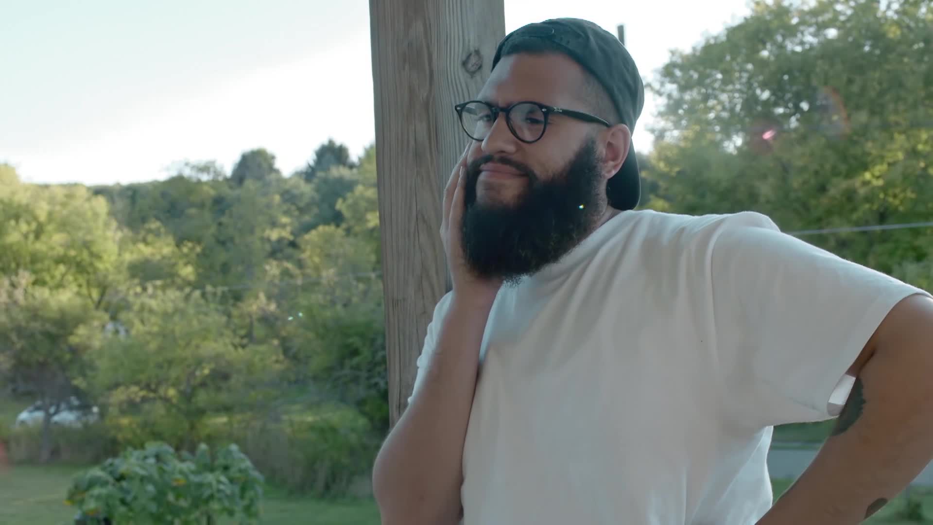 A man with a full beard and glasses leans against a wooden post, his hand resting on his cheek. He's wearing a baseball cap and a white t-shirt, with trees and sunlight in the background.
