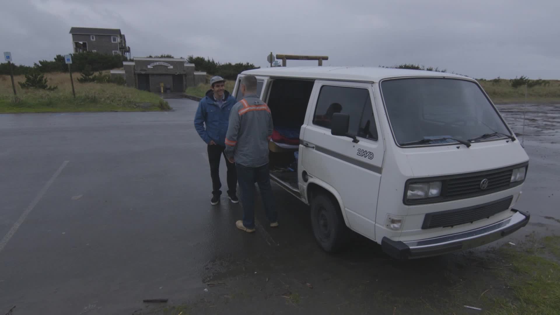 Two men stand by the open door of a white van, talking in a wet parking lot. The overcast sky and the building in the background suggest a coastal location.
