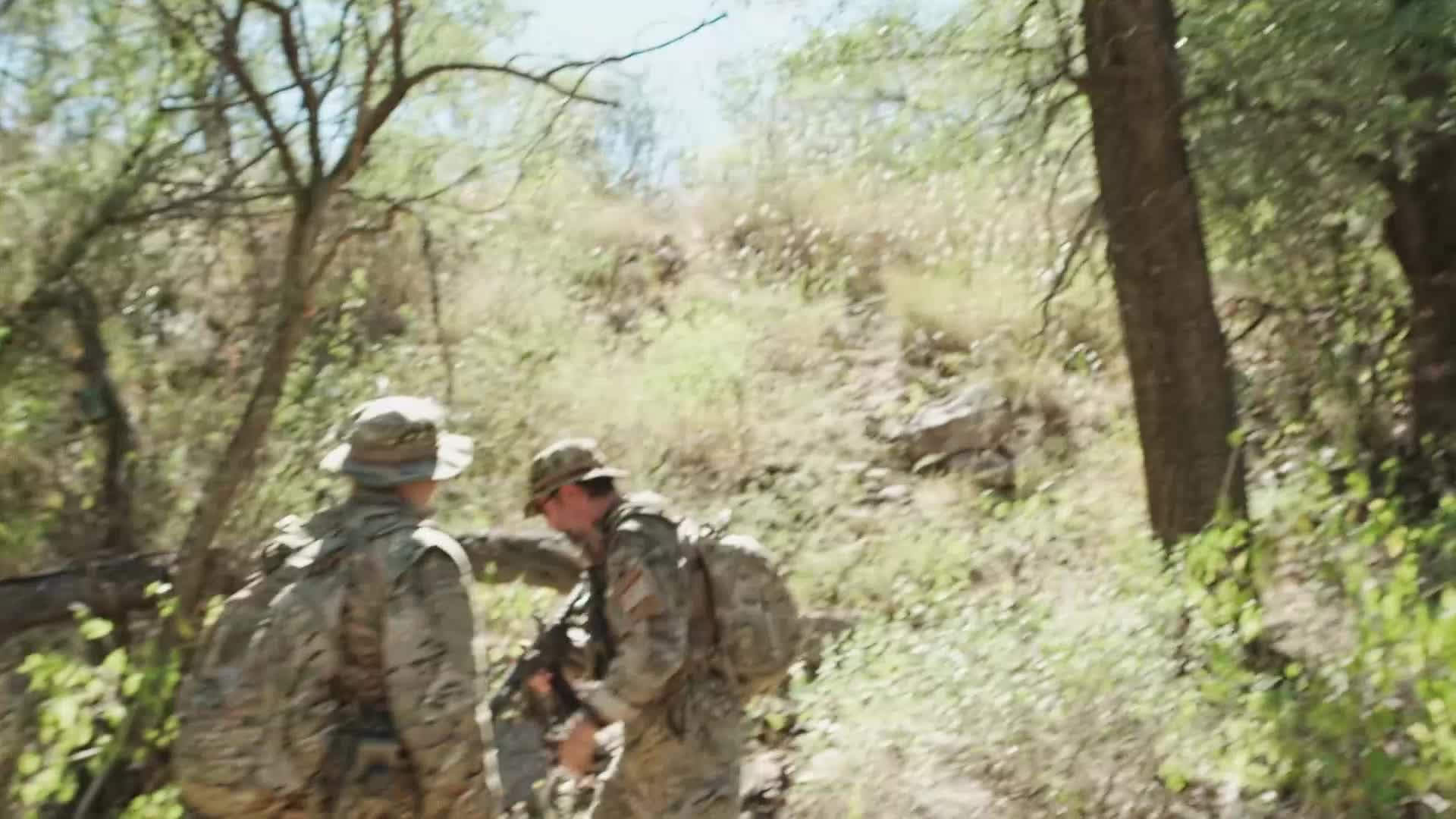 Two soldiers in camouflage gear are hiking up a rocky, wooded slope. One points ahead, while the other carries a rifle.
