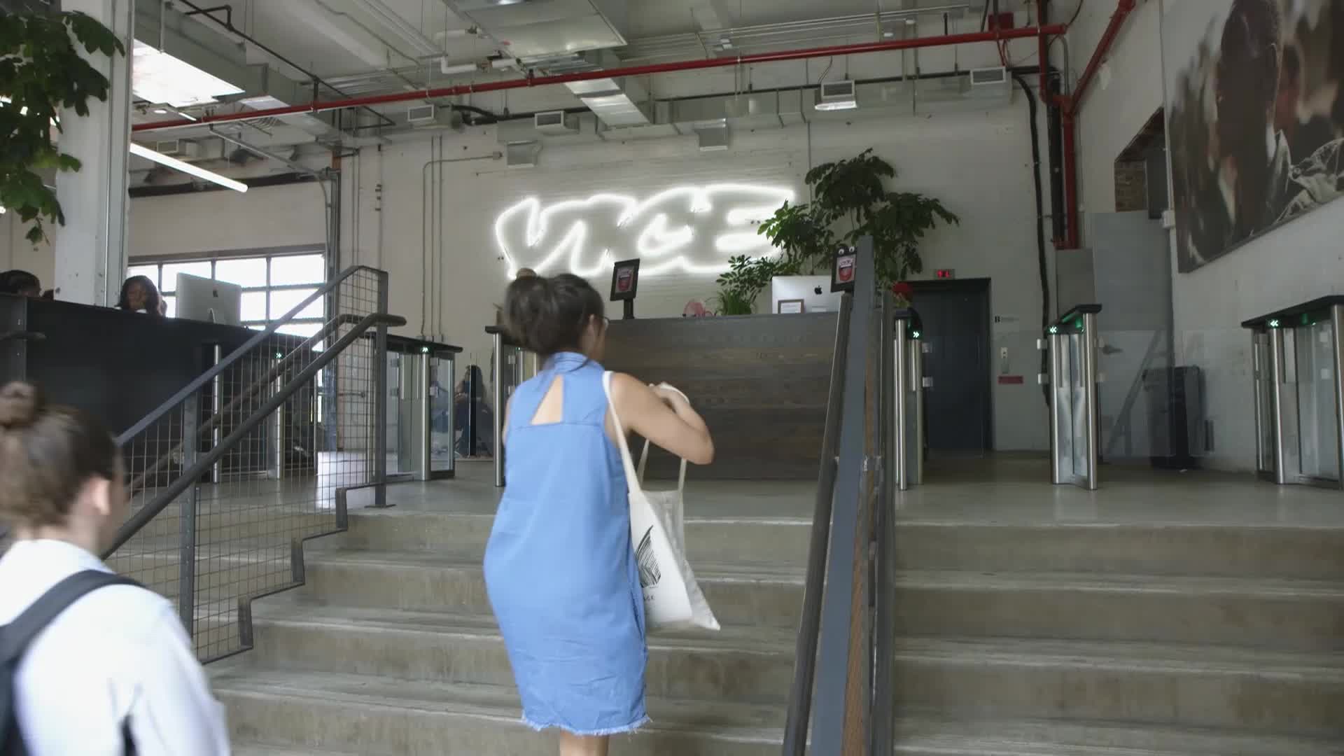 People are ascending a concrete staircase toward the VICE logo, a bright white neon sign. A woman in a blue dress carries a tote bag, heading toward the reception desk.
