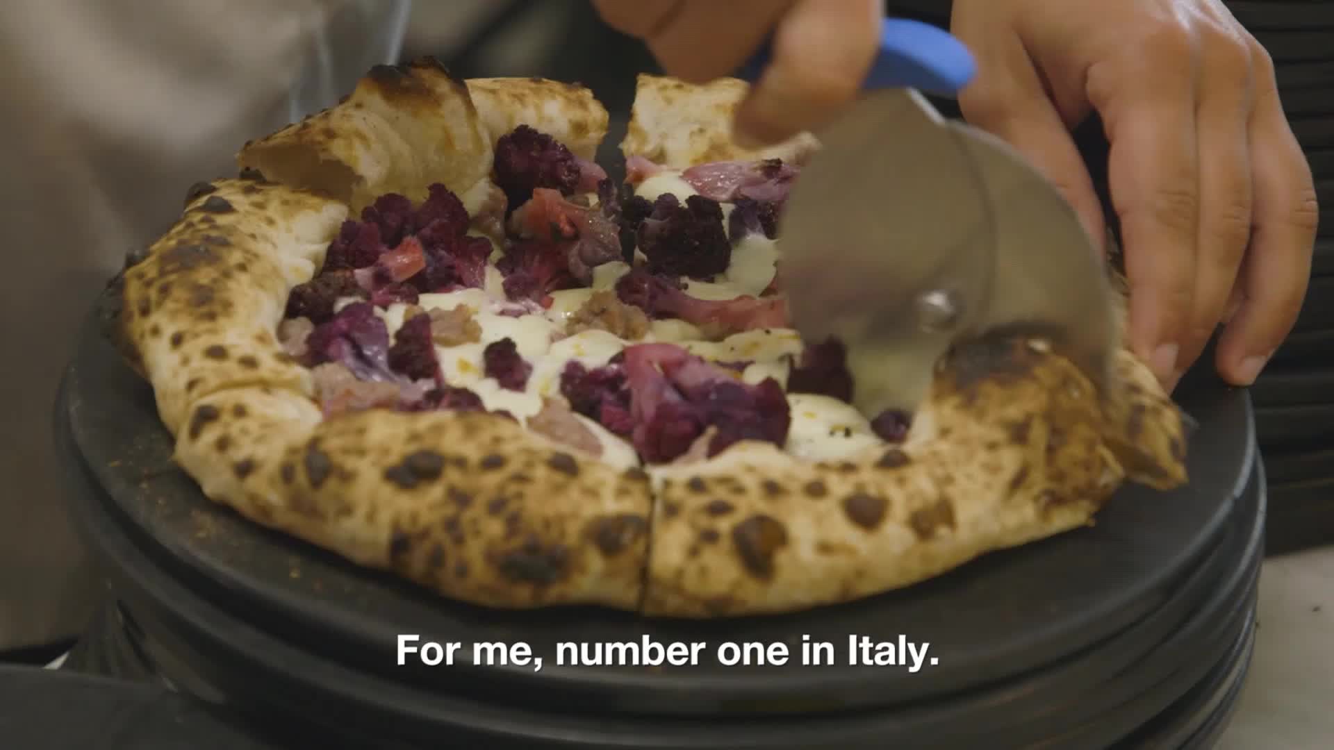 A pizza cutter slices through a pizza topped with purple vegetables and white cheese. The crust is dark and bubbly, and the pizza sits on a dark, round surface.
