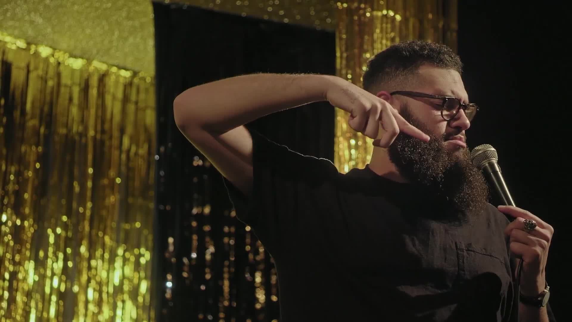 The man on stage is pointing at his beard with a serious expression. He's holding a microphone, ready to speak to the audience.
