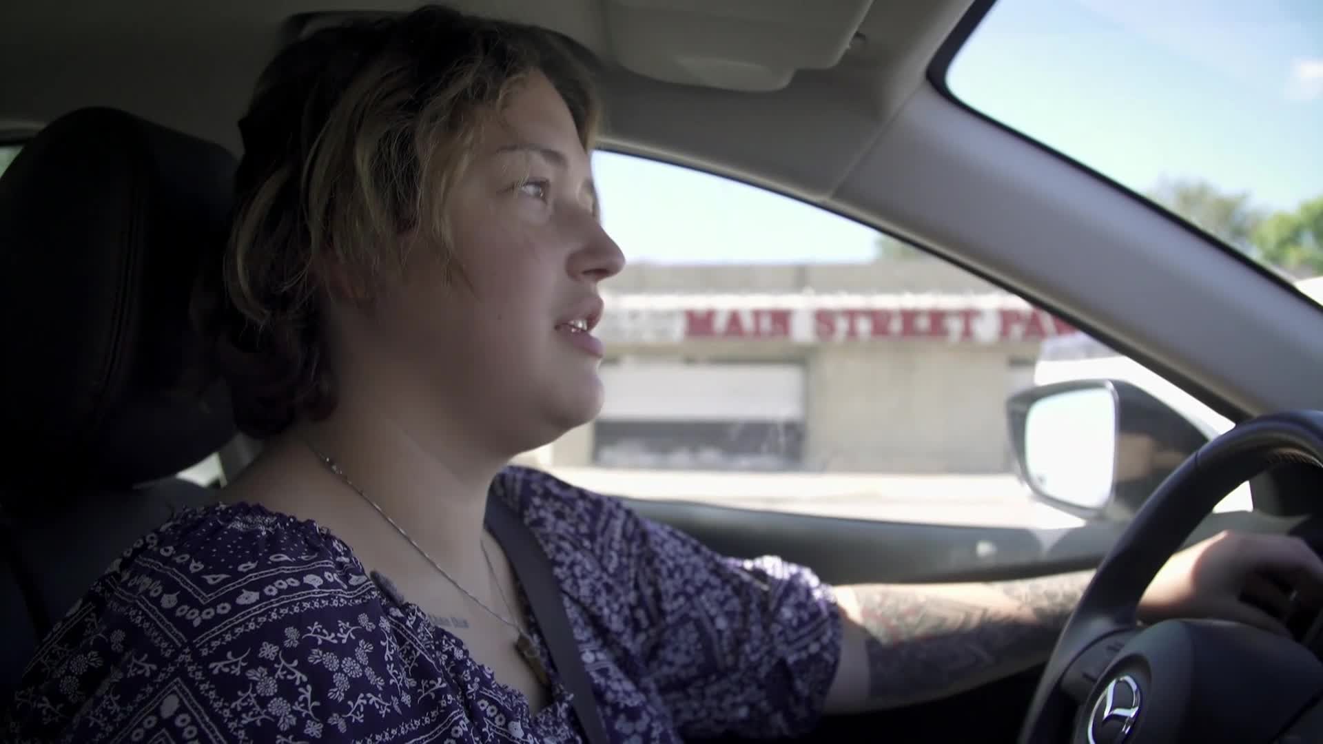 A woman with short, light-colored hair drives a car, speaking, her gaze directed off-screen. The "Main Street Park" sign is visible through the windshield, suggesting a location in the United States.
